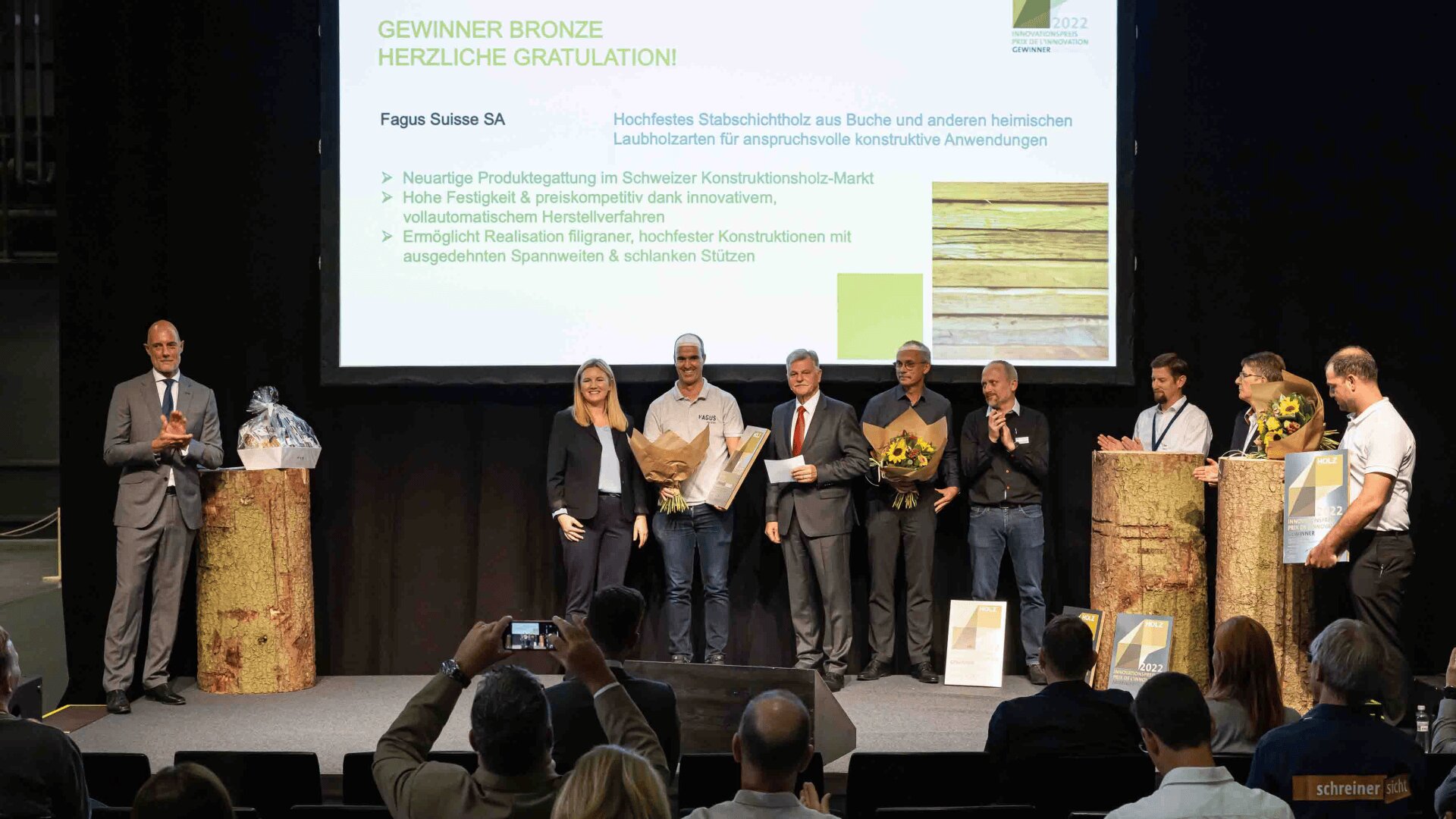 Eine Gruppe von Personen auf der Bühne einer Preisverleihung posiert mit Blumensträußen und Urkunden, während auf einem Bildschirm hinter ihnen die Fagus Suisse SA als Bronzegewinnerin bekannt gegeben wird, wobei Text und Bilder von Holzplatten angezeigt werden.