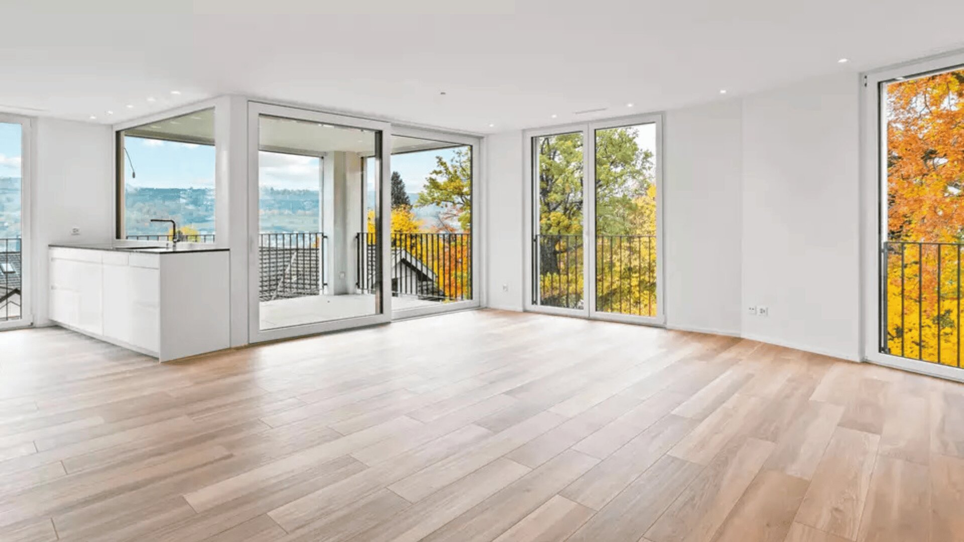 Eine geräumige, moderne Wohnung mit hellem Holzfußboden, großen Fenstern und Glastüren, die zu Balkonen führen. Der Raum ist hell und bietet einen Blick auf die herbstlichen Bäume und einen entfernten Hügel. Auf der linken Seite ist eine weiße Küche zu sehen.