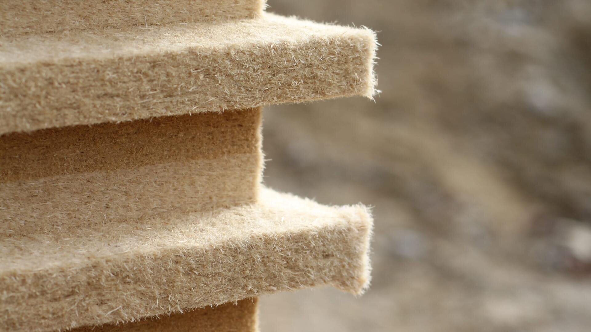 Nahaufnahme der rauen, faserigen Kante einer geschichteten, beigen Dämmplatte, die wahrscheinlich aus natürlichen Materialien besteht, vor einem unscharfen braunen Hintergrund.