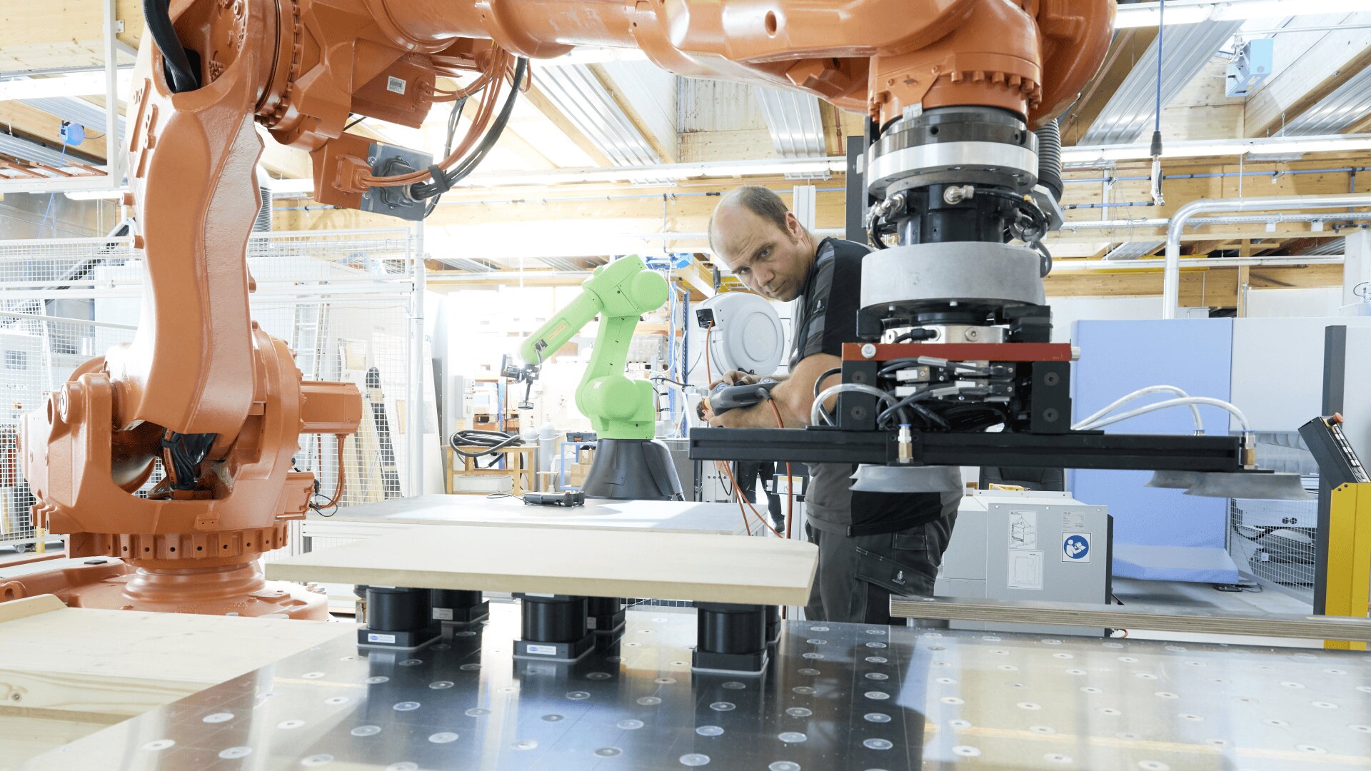 Ein Mann bedient einen großen orangefarbenen Industrieroboterarm in einer modernen Werkstatt, während im Hintergrund ein weiterer grüner Roboterarm und verschiedene Geräte zu sehen sind.