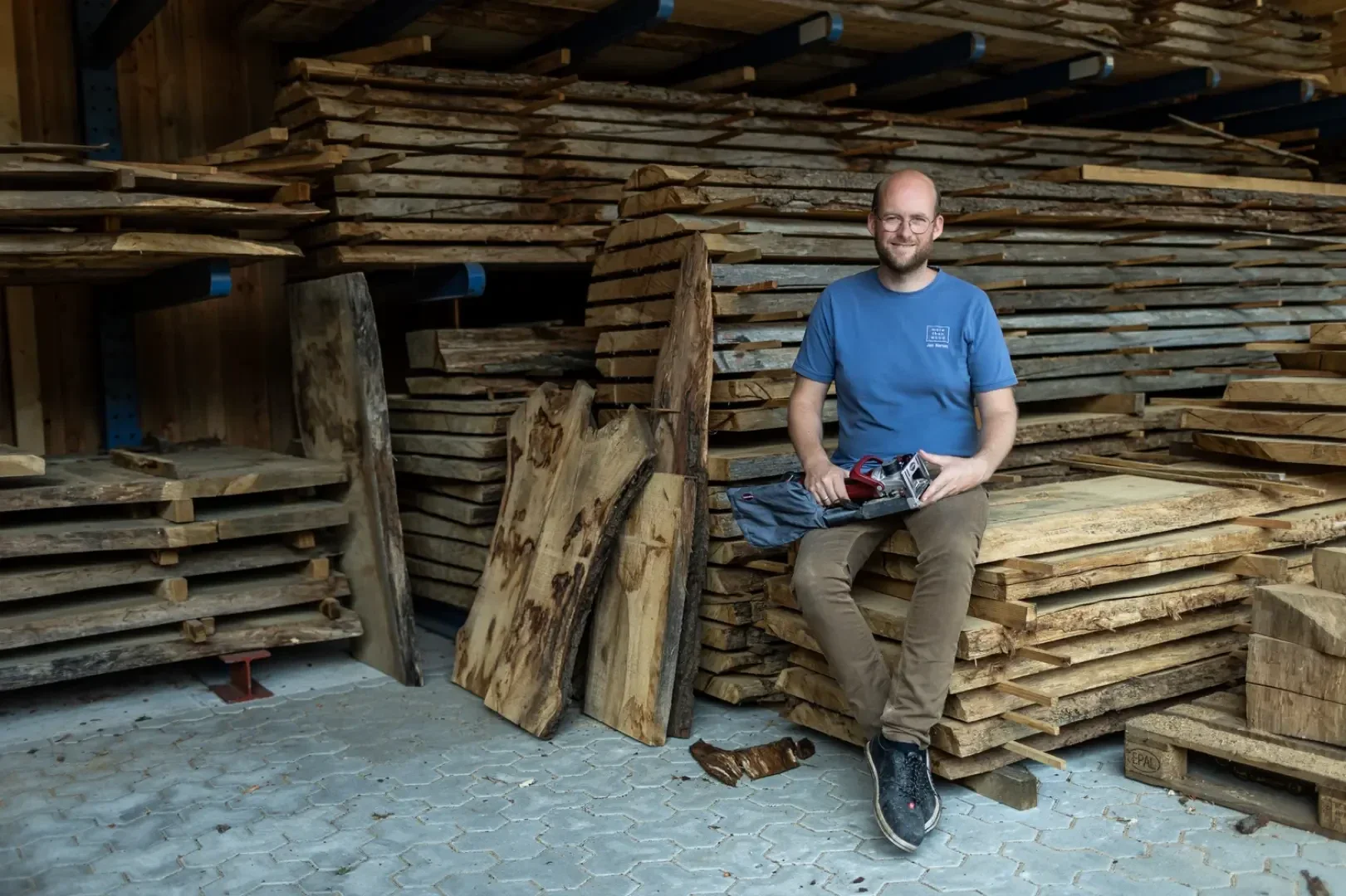 Un homme souriant, vêtu d'une chemise bleue et d'un pantalon marron, est assis sur des planches de bois empilées dans un entrepôt de bois, un outil électrique à la main. D'autres planches de bois brut sont visibles autour de lui.