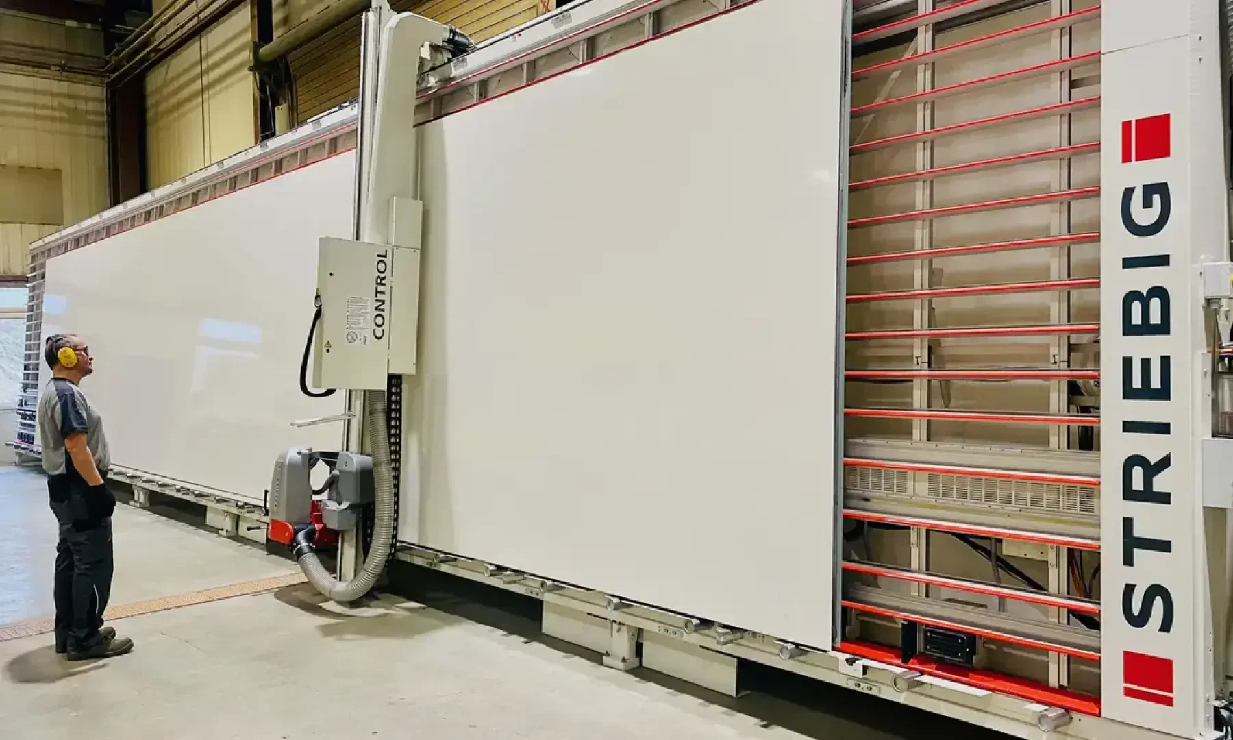 Eine Person mit Gehörschutz steht vor einer großen, vertikalen STRIEBIG-Plattensägemaschine in einer Industrieumgebung, auf der eine weiße Platte zum Schneiden montiert ist.