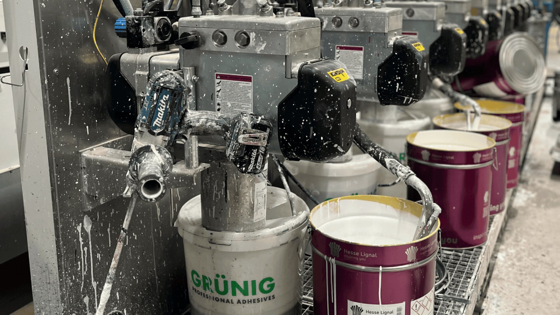 Des machines automatisées distribuent de l'adhésif blanc dans des seaux ouverts, dont un seau blanc étiqueté GRÜNIG et des conteneurs violets. La zone de travail présente des éclaboussures de colle et des équipements industriels en cours d'utilisation.