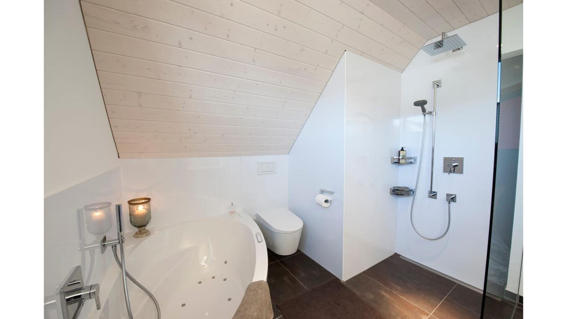 Salle de bains moderne avec un plafond en bois incliné, comprenant une baignoire blanche avec des bougies, des toilettes murales, un porte-papier hygiénique et une douche vitrée avec des étagères et un pommeau de douche à effet de pluie.
