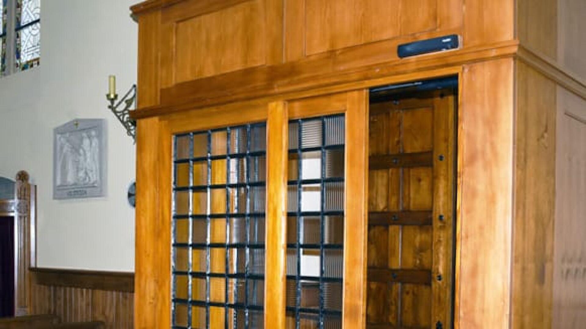 Eine hölzerne Beichtkabine mit schwarzen Metallgittern und Schiebetüren befindet sich in einer Kirche, in der im Hintergrund Buntglasfenster und eine Wandtafel zu sehen sind.
