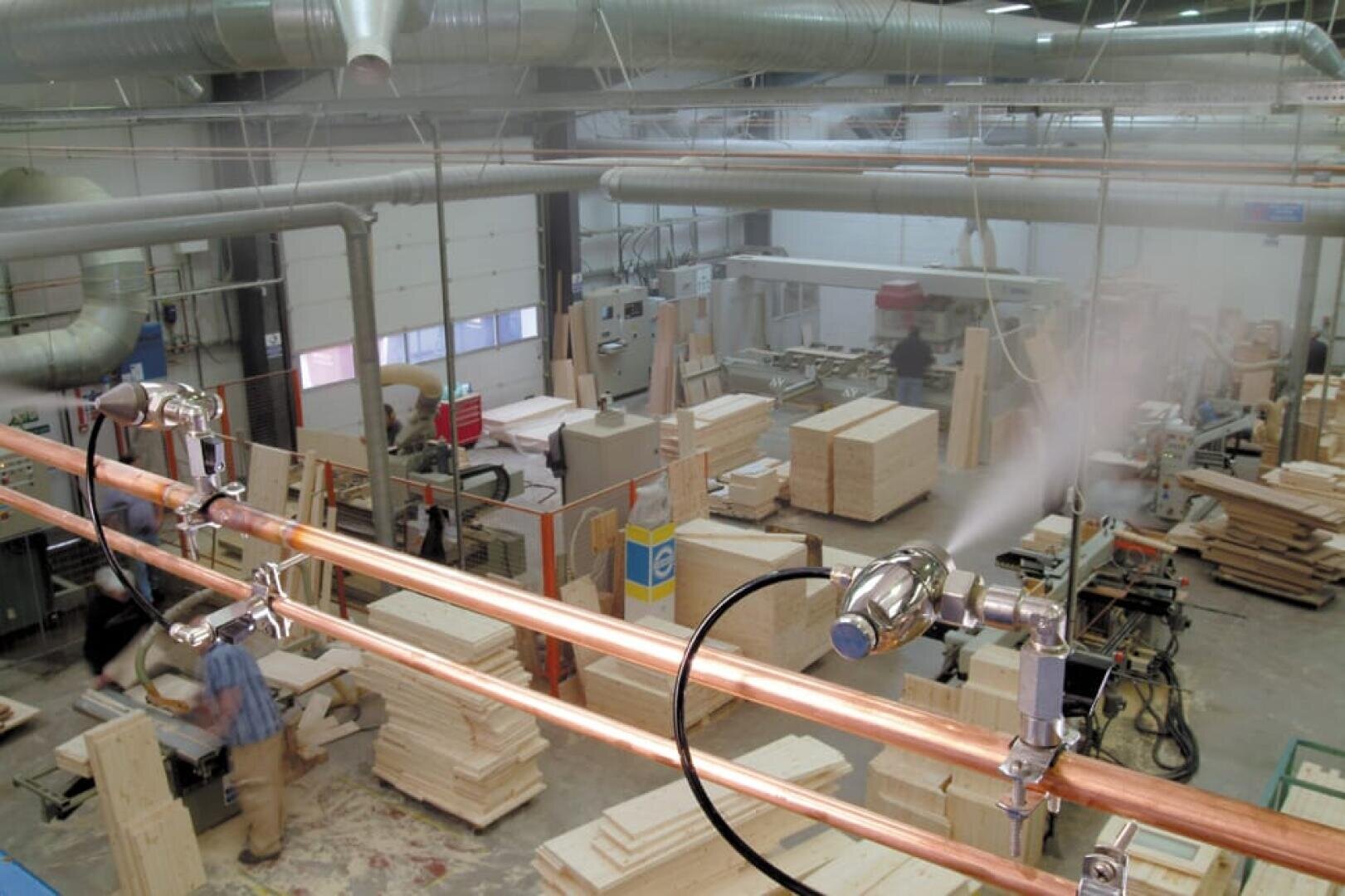 Eine große Holzverarbeitungsfabrik mit Stapeln von unfertigen Holzkisten, verschiedenen Werkzeugen und Geräten sowie Arbeitern, die Produkte zusammensetzen. Über den Köpfen versprüht ein Vernebelungssystem Wasserdampf, um die Luftqualität und den Staub zu kontrollieren.