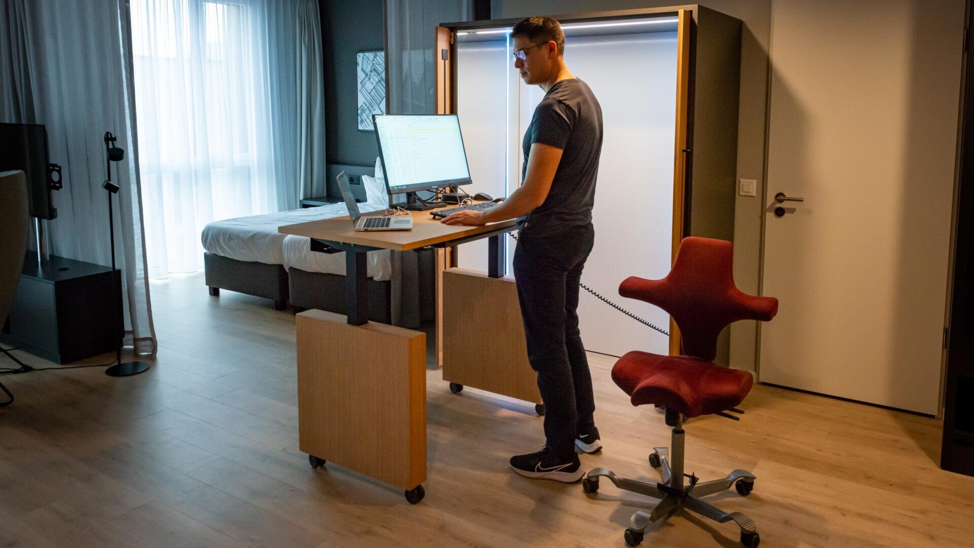 Ein Mann steht und arbeitet an einem höhenverstellbaren Schreibtisch mit einem Computer in einem modernen, gut beleuchteten Raum. In der Nähe steht ein einzigartiger roter ergonomischer Stuhl, und im Hintergrund ist ein Bett zu sehen.