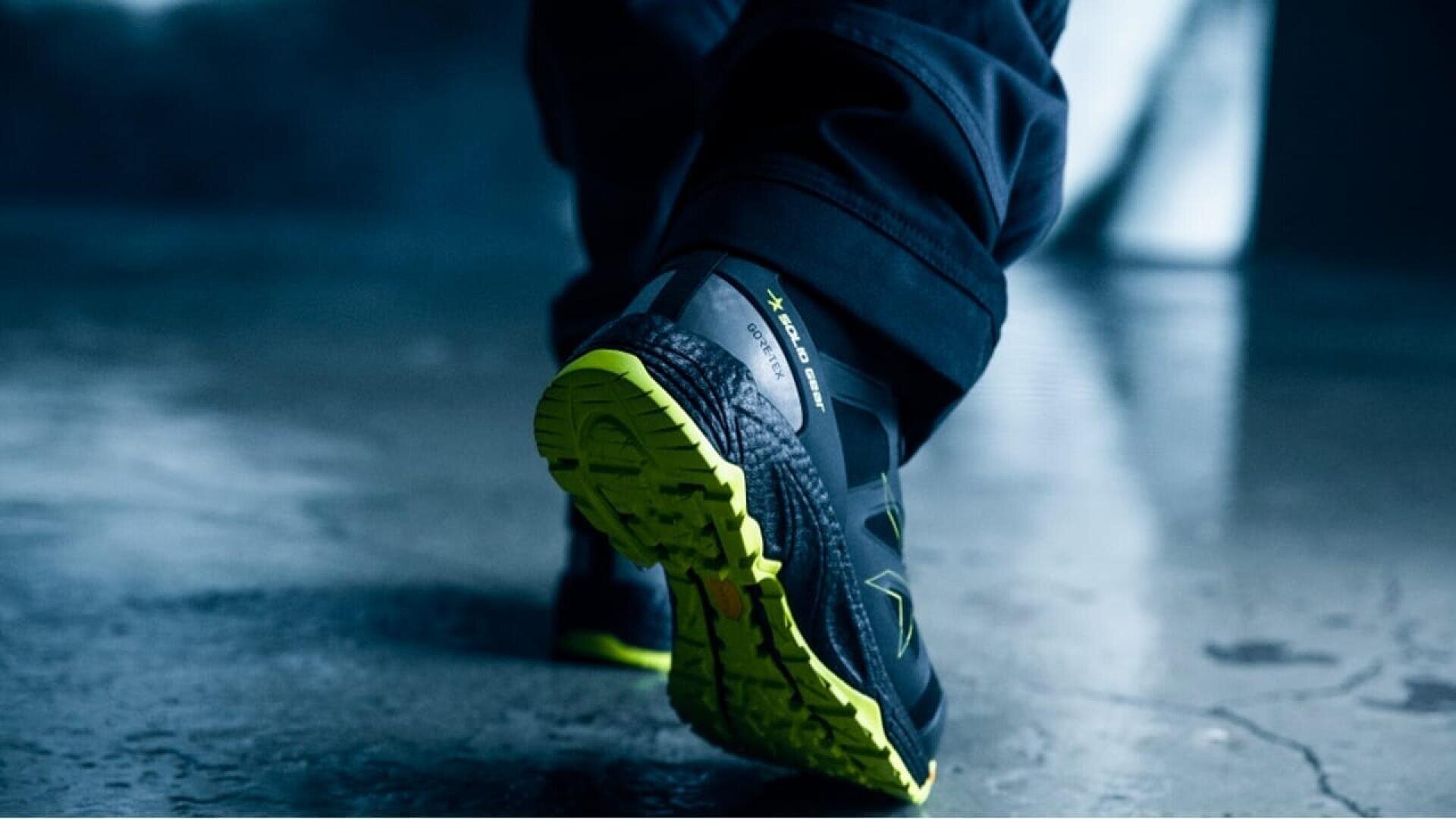 Gros plan d'une personne portant des chaussures de sport noires et vertes fluo, marchant sur une surface humide et réfléchissante. L'accent est mis sur la semelle de la chaussure lorsque la personne fait un pas en avant.