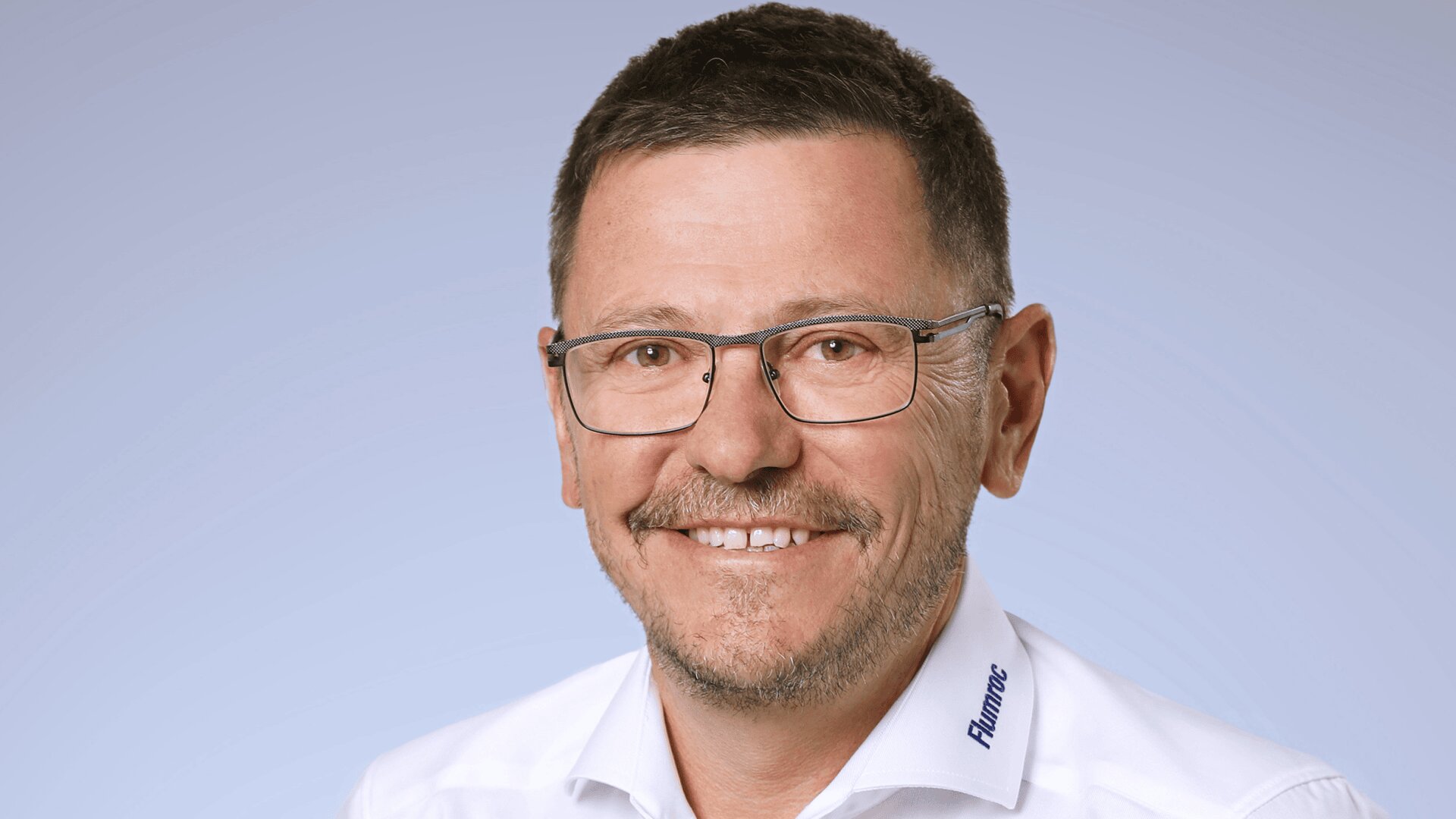 Un homme aux cheveux bruns courts, aux lunettes et à la barbe taillée sourit à l'appareil photo. Il porte une chemise blanche avec l'inscription Aumate sur le col, sur un fond bleu clair.