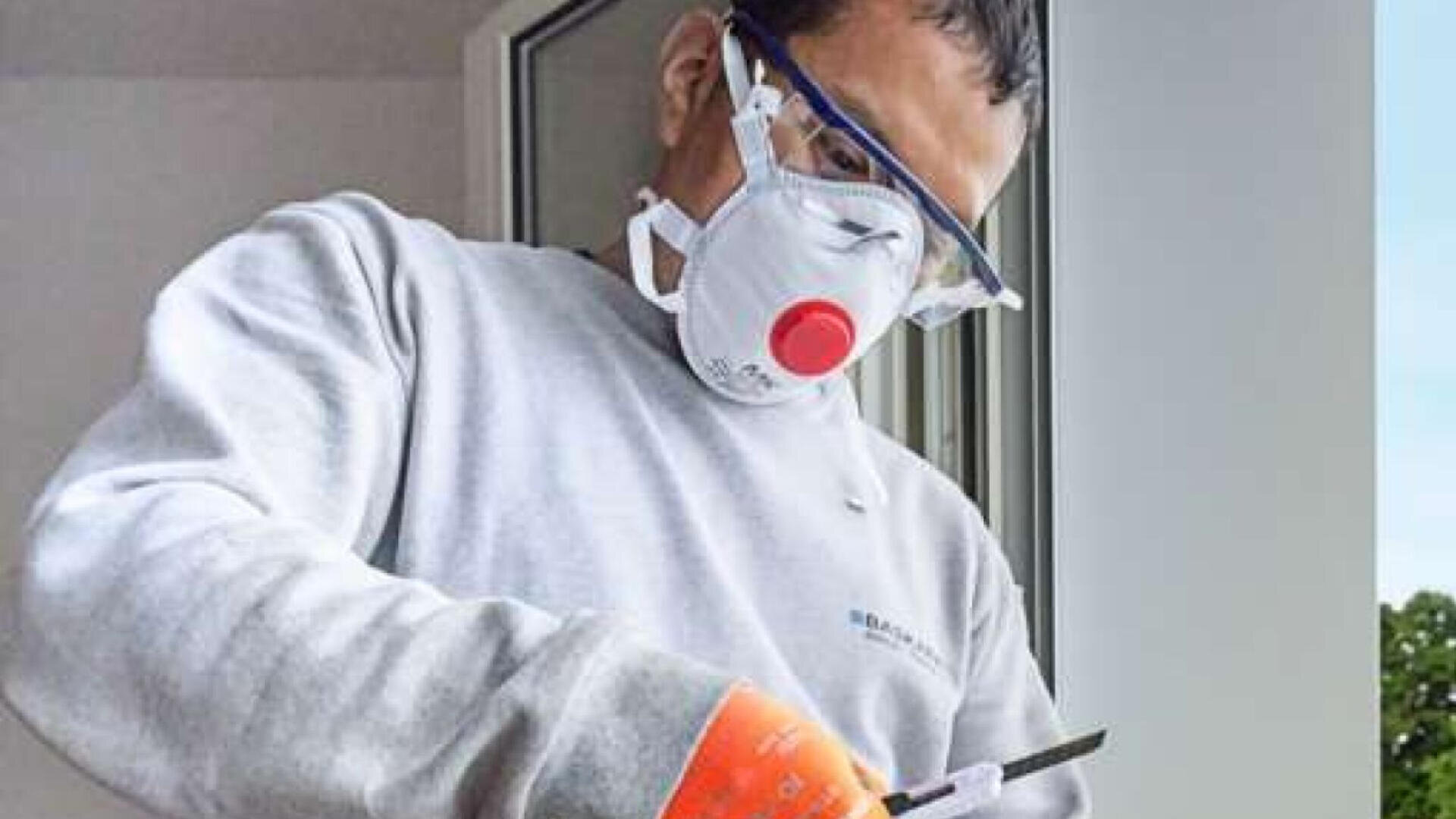 Eine Person, die eine Schutzbrille, eine N95-Maske, Handschuhe und ein graues Sweatshirt trägt, schneidet in einem Raum in der Nähe eines offenen Fensters ein Material zu.
