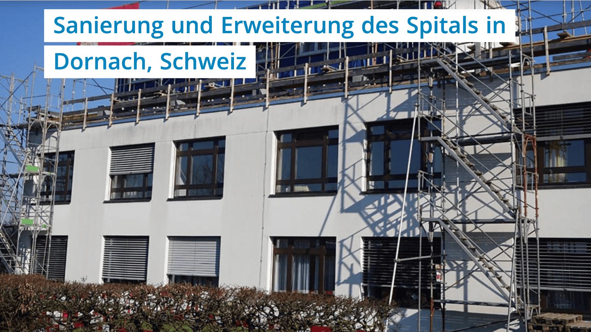 Ein weißes Spitalgebäude in Dornach, Schweiz, das renoviert und erweitert wird, mit einem Gerüst an der Außenseite und einem Schild mit der Aufschrift Sanierung und Erweiterung des Spitals in Dornach, Schweiz.