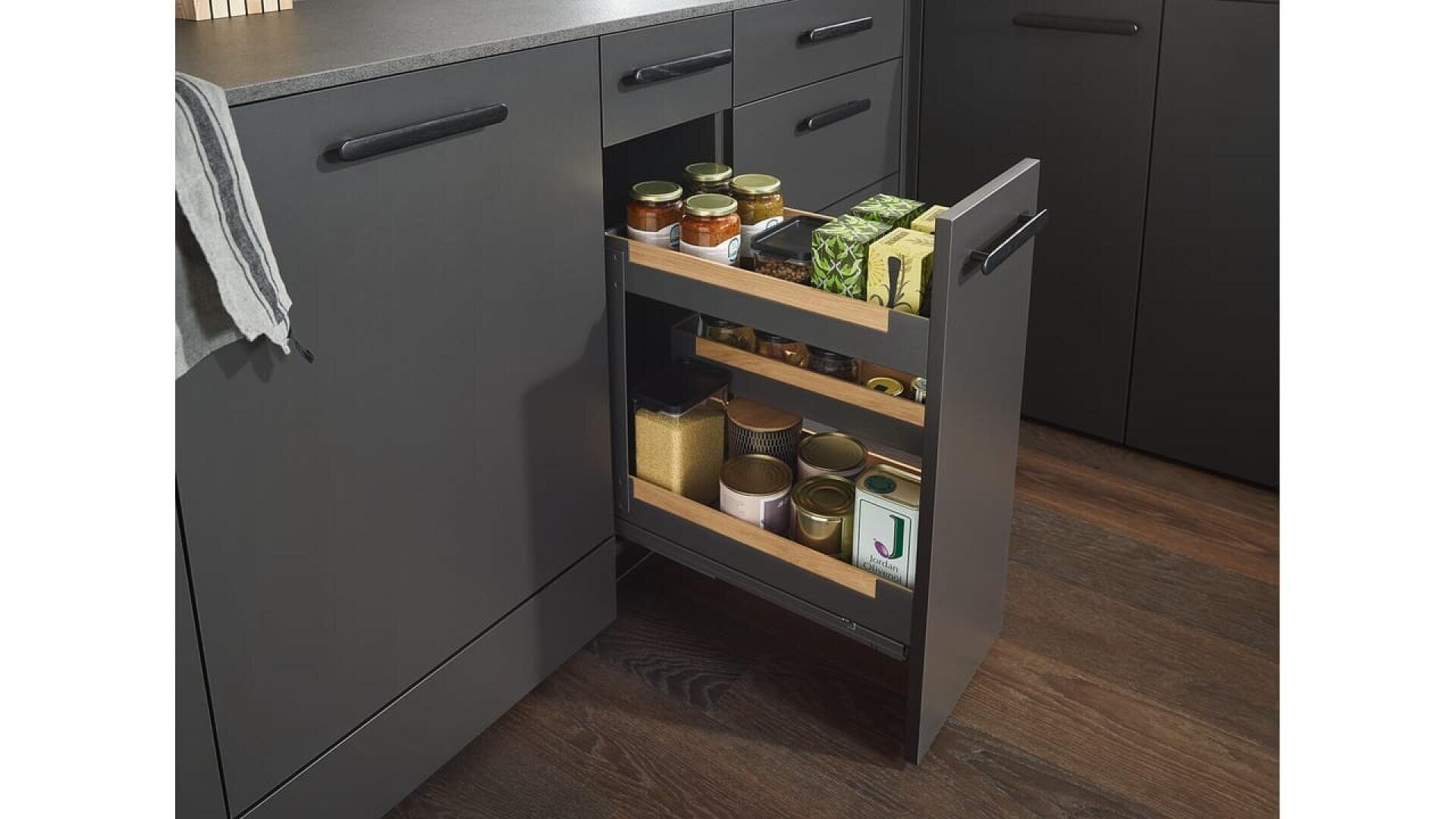 Une cuisine moderne et élégante avec un tiroir d'armoire sombre ouvert, révélant des bocaux, des boîtes de conserve et des paquets d'aliments bien rangés sur deux étagères. Les armoires et le sol sont de couleur foncée.