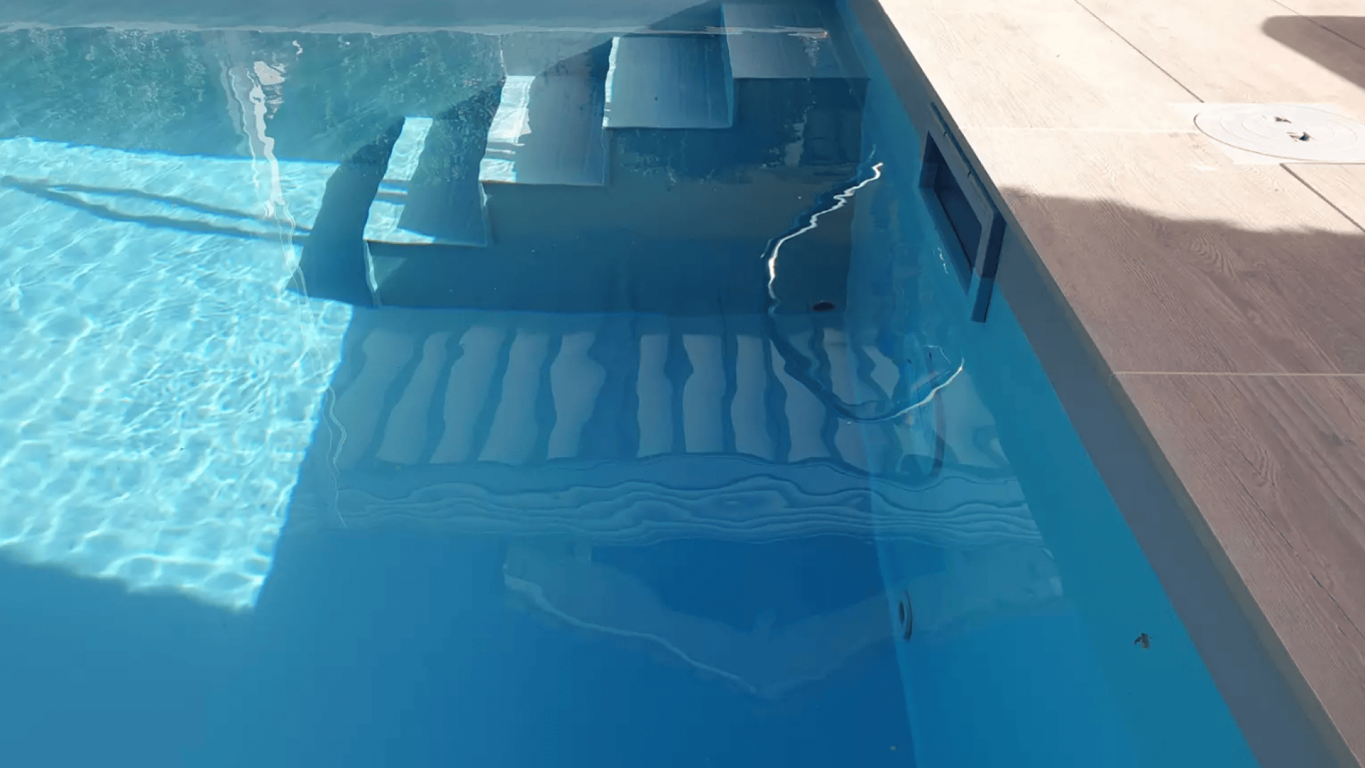 Ein Pool mit Stufen unter Wasser und der Spiegelung eines Schwimmers auf der Wasseroberfläche. Das Sonnenlicht erzeugt Muster im hellblauen Wasser, und der Rand des Pools ist rechts zu sehen.