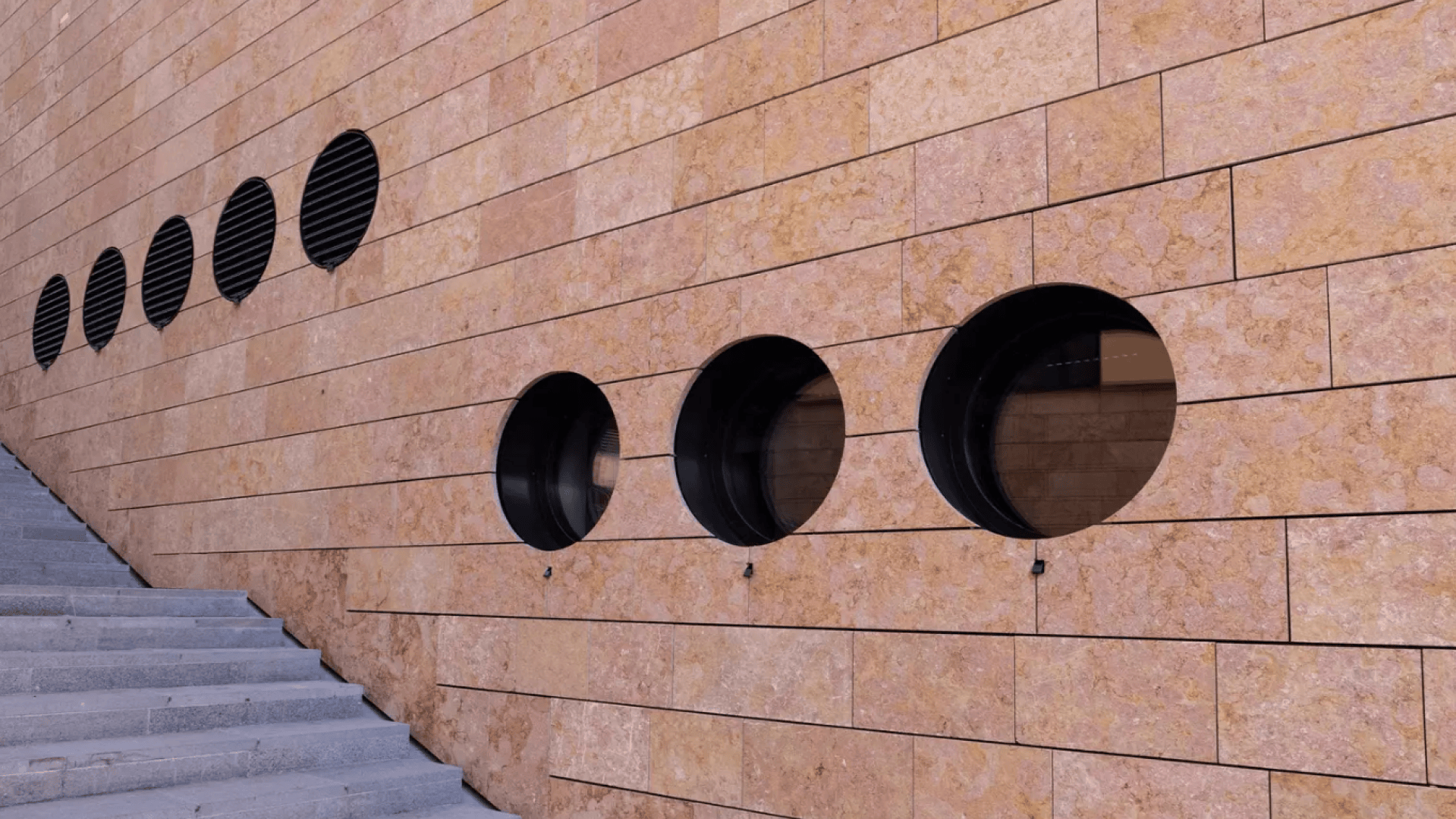 Un mur en pierre de couleur havane avec cinq fenêtres circulaires bordées de noir en rangée, à côté d'un escalier aux marches gris clair, créant un motif architectural moderne.