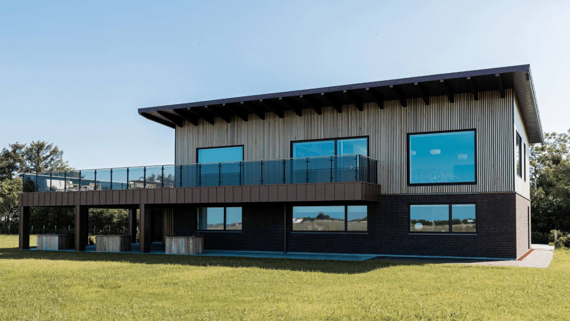 Modernes zweistöckiges Haus mit grossen Glasfenstern, einem geräumigen Balkon mit Glasgeländer und einer Mischung aus Holz- und dunklen Ziegelfassaden, umgeben von grünem Gras und Bäumen unter einem klaren blauen Himmel. Augenmerk ist auf die Holzfassade von Aluwood im Panel-Look.