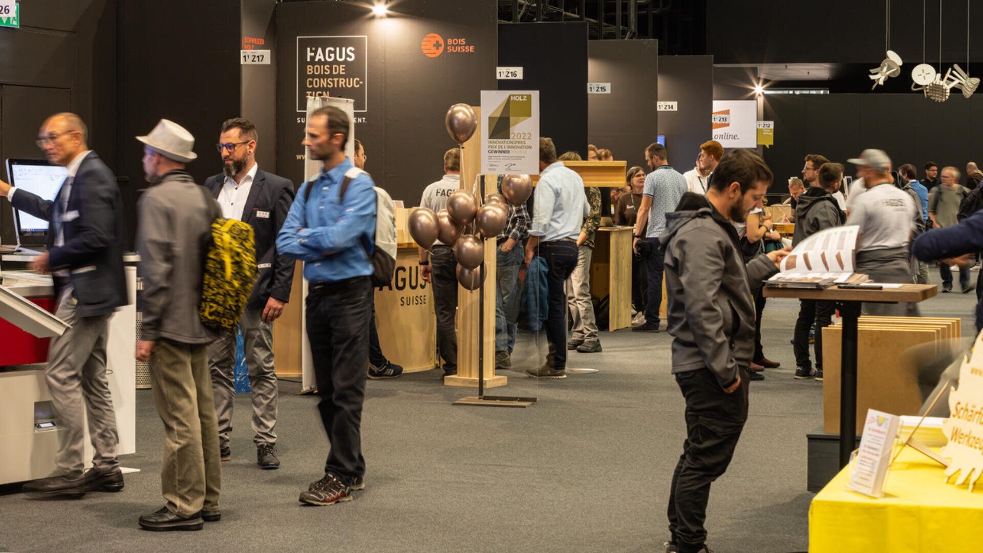 Auf einer Indoor-Messe oder -Ausstellung gehen Menschen umher und interagieren. Verschiedene Stände mit Informationsdisplays, Schildern und Luftballons sind zu sehen, und die Besucher sind in Gespräche vertieft oder erkunden die Ausstellungsstücke.