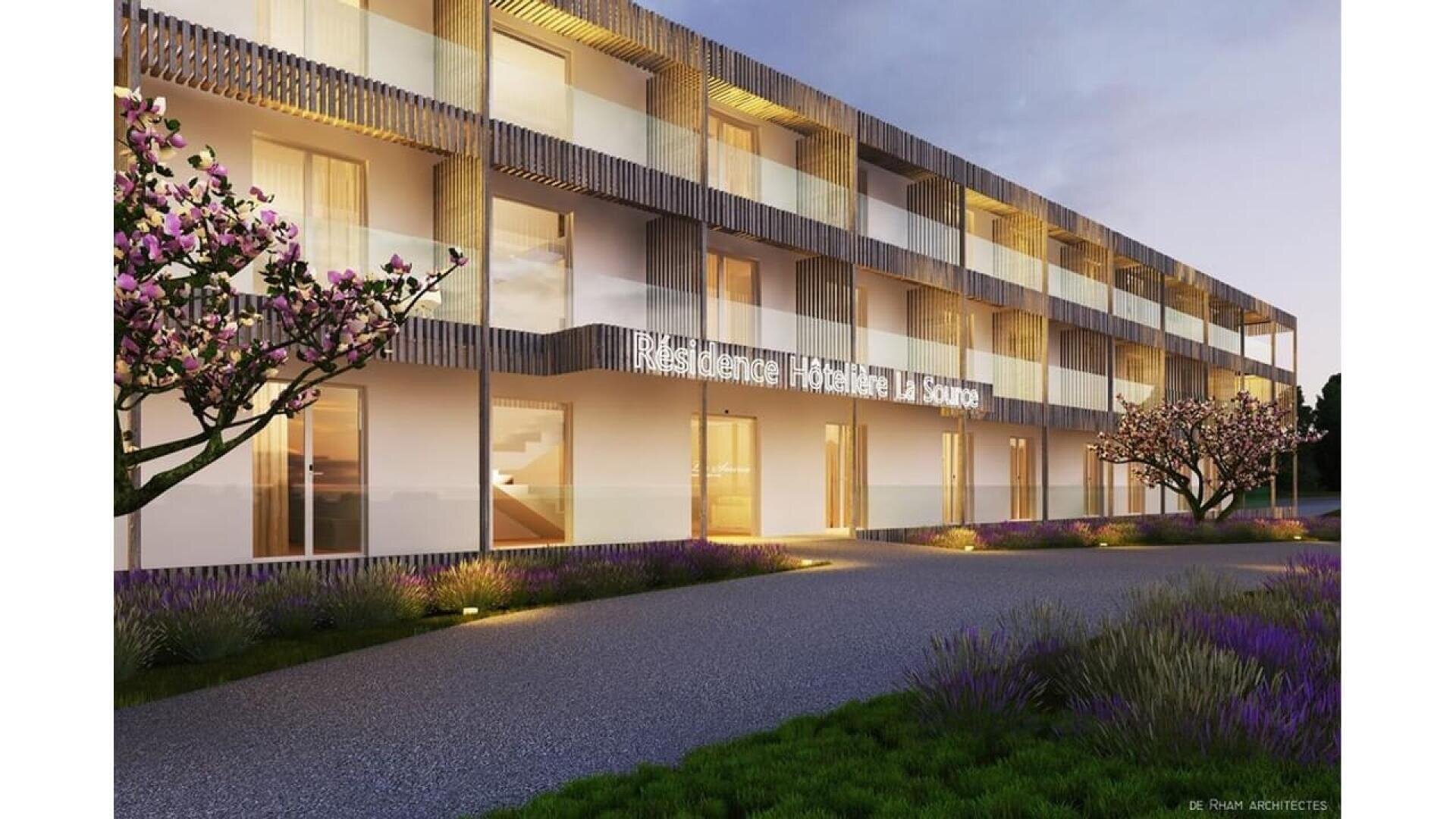 La Résidence Hôtelière La Source est un bâtiment moderne de trois étages, entouré de jardins paysagers avec des arbres en fleurs et éclairé par des lumières extérieures chaleureuses à la tombée de la nuit.