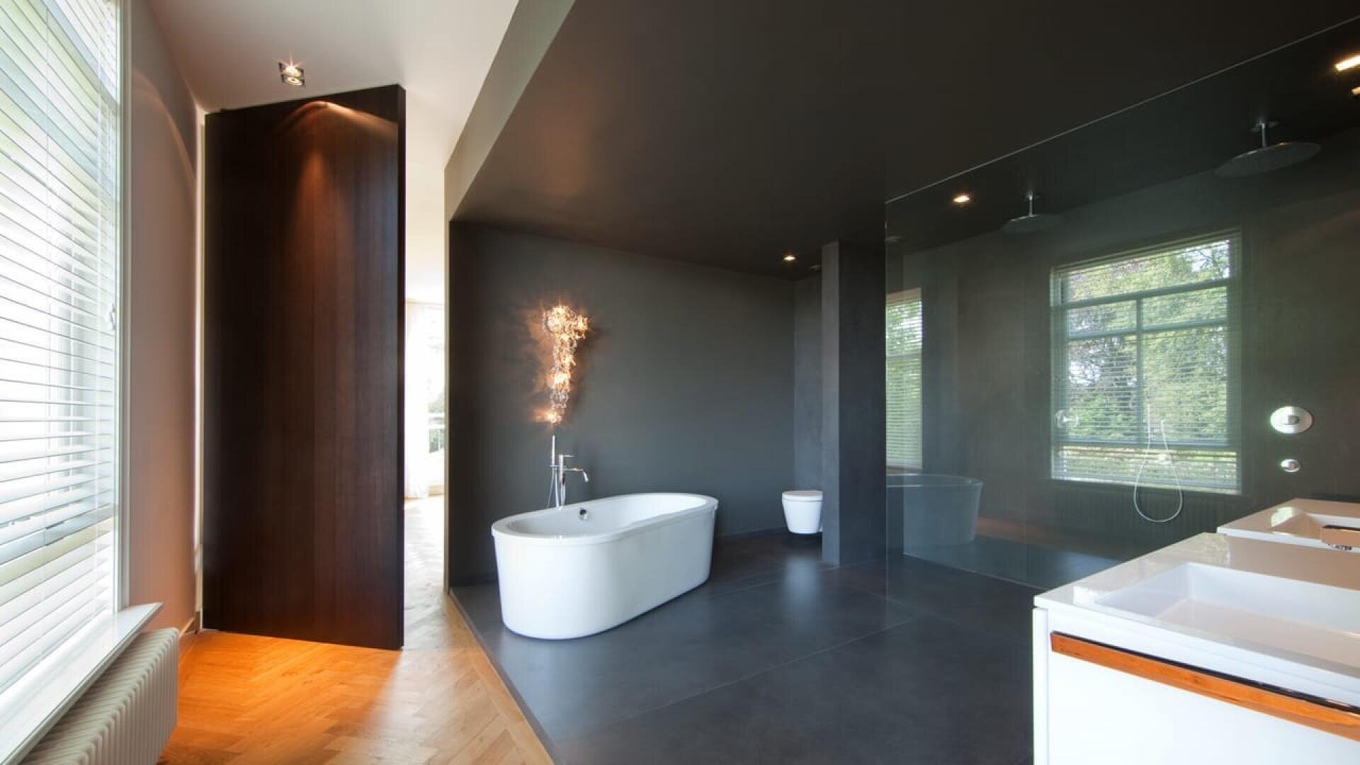 Salle de bains moderne et minimaliste avec une baignoire blanche sur pied, des murs et un sol sombres, une grande douche en verre, des lavabos blancs, des toilettes et une grande fenêtre laissant entrer la lumière naturelle.
