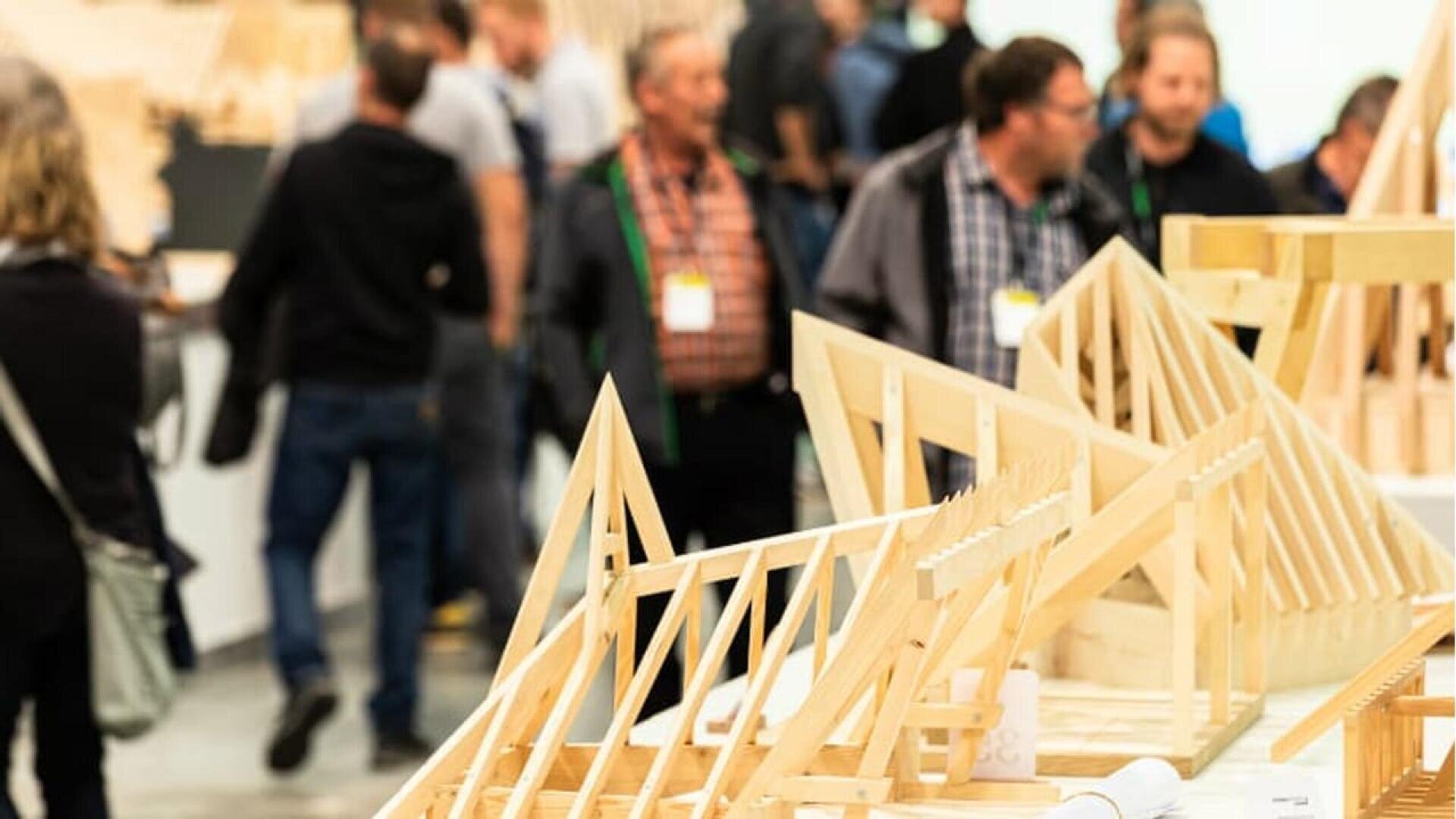 Des modèles architecturaux en bois de fermes de toit sont exposés sur des tables au premier plan. À l'arrière-plan, un groupe de personnes est rassemblé, conversant et observant les modèles lors de ce qui semble être une exposition ou une conférence.
