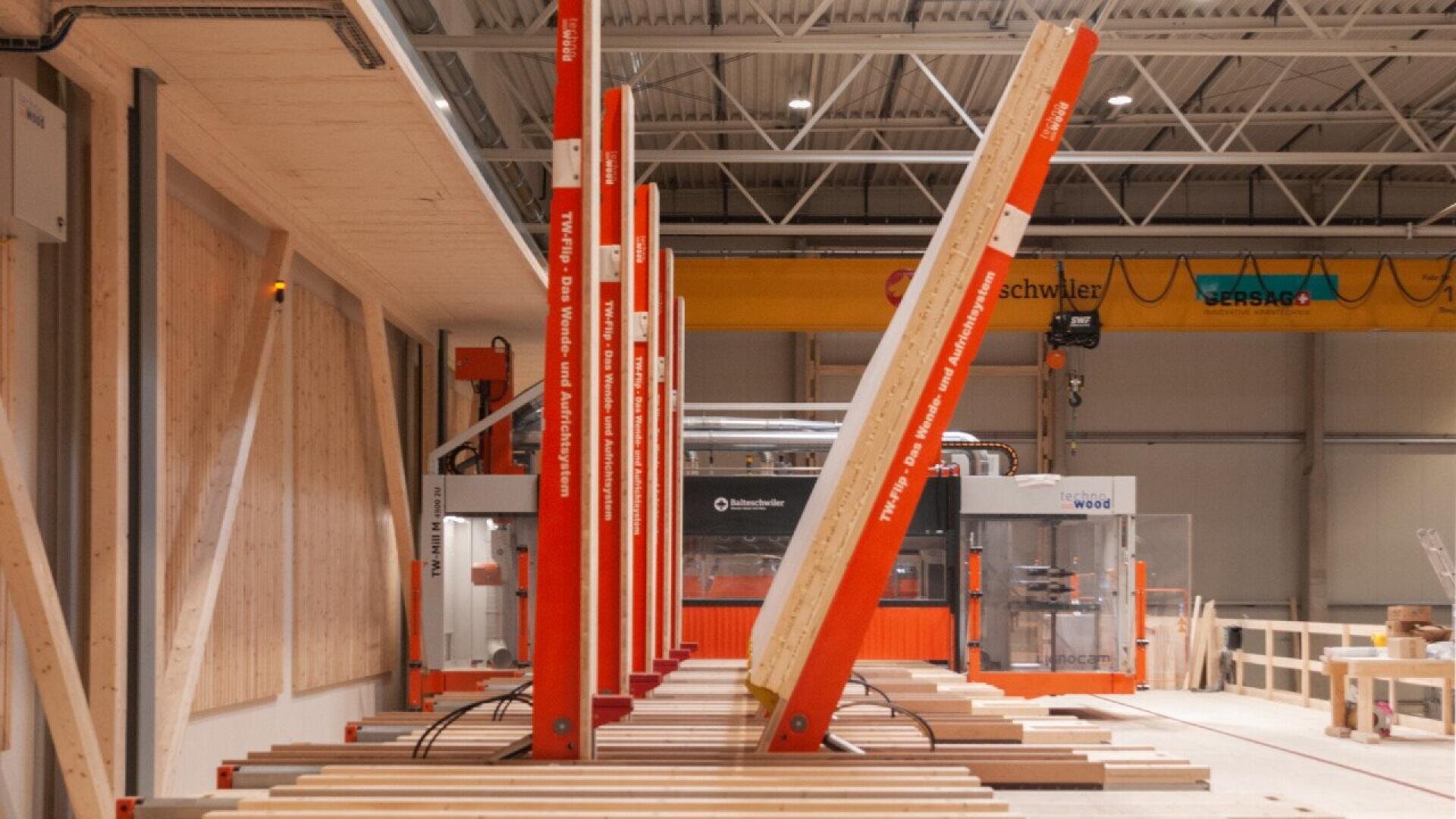 Rangées de grandes poutres en bois maintenues debout par des supports métalliques rouges dans un atelier industriel moderne et bien éclairé, avec des machines et des outils en arrière-plan.