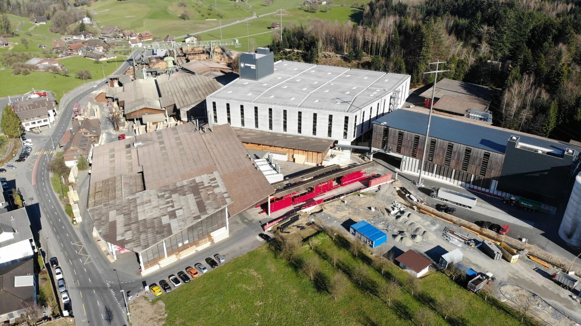 Luftaufnahme einer Industrieanlage mit mehreren großen Gebäuden, Baustellen, parkenden Autos und nahe gelegenen Straßen, umgeben von grünen Feldern, Bäumen und einem kleinen Dorf.