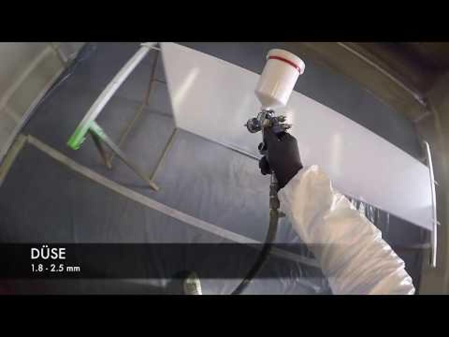 Une personne portant des vêtements de protection et des gants utilise un pistolet pour appliquer de la peinture ou un revêtement sur une surface blanche ; le texte sur l'image indique "DÜSE 1.8 - 2.5 mm.".