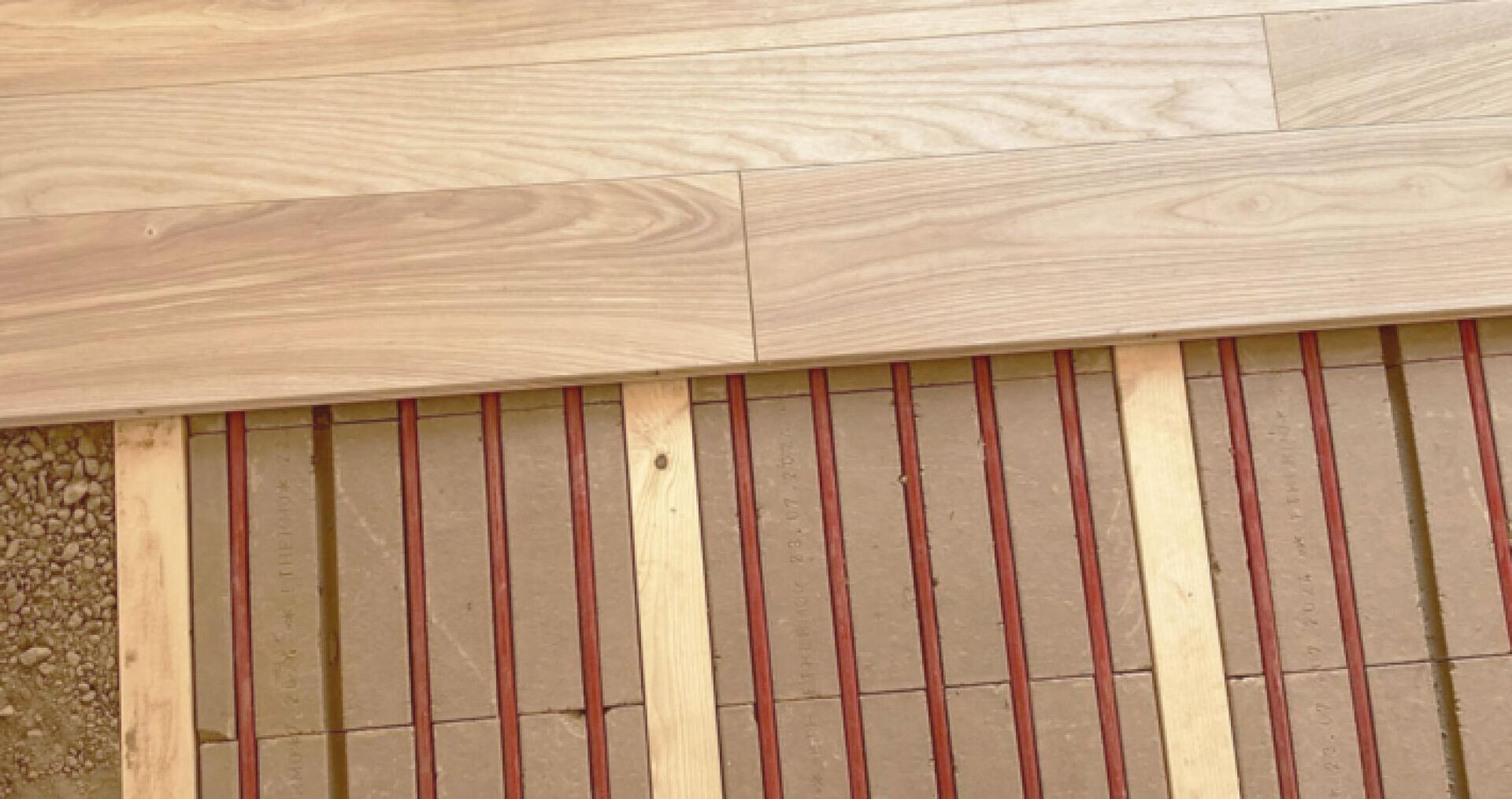 Teilweise verlegter Parkettboden mit Holzdielen auf der Oberseite und freiliegendem Fußbodenheizungssystem mit roten Heizelementen und Holzlatten darunter.