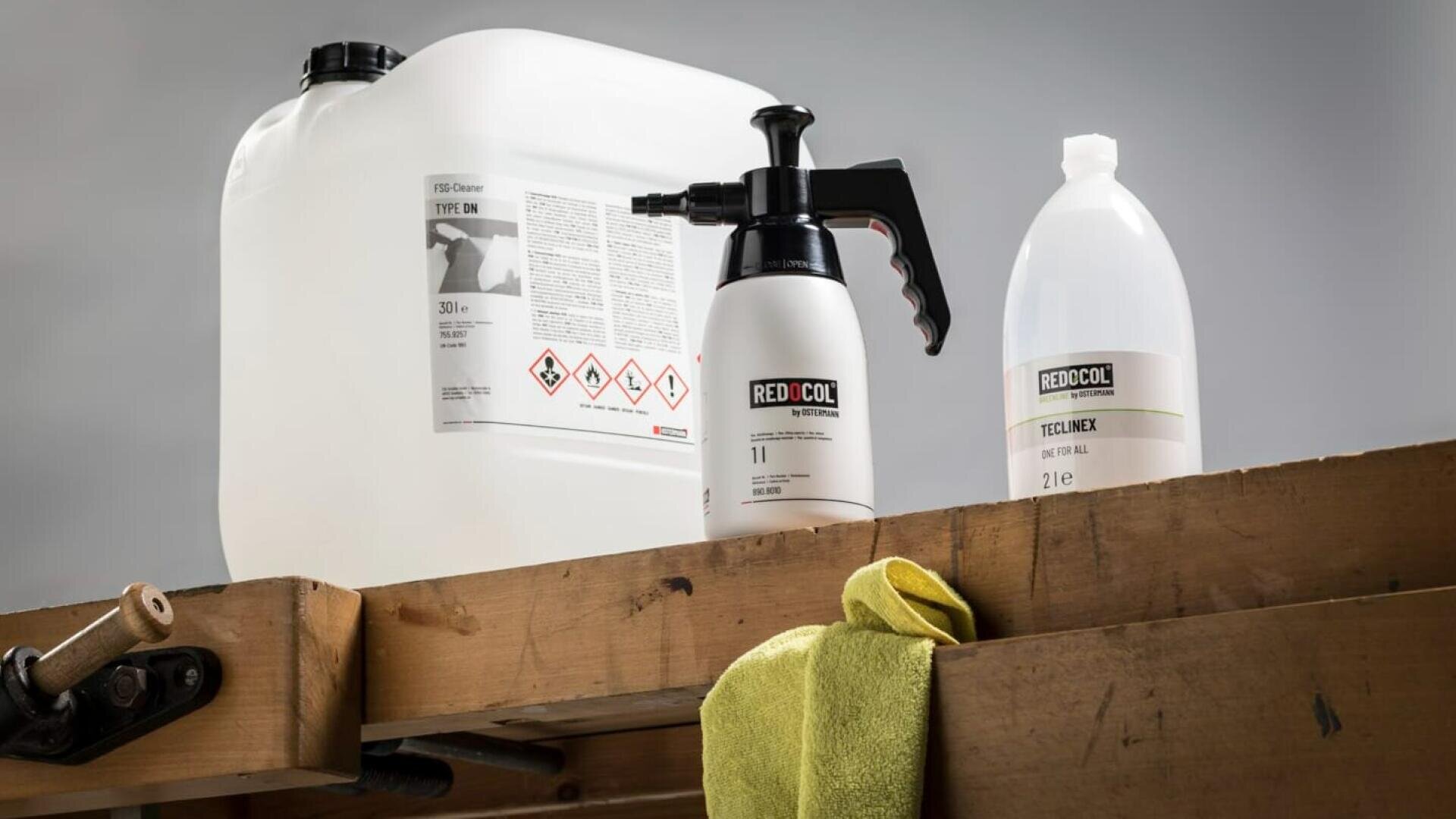 Un grand récipient en plastique blanc, deux flacons pulvérisateurs étiquetés RED COOL et un flacon transparent sont posés sur une surface en bois avec un chiffon jaune drapé sur le bord. L'arrière-plan est gris neutre.