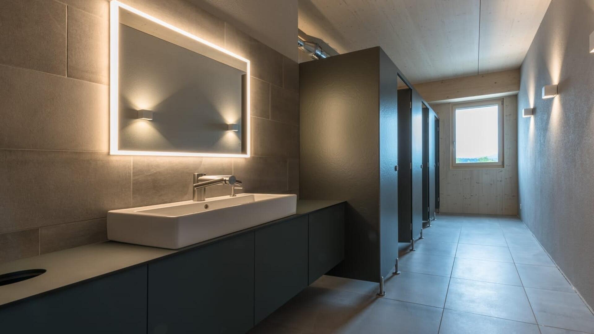 Toilettes publiques modernes avec un grand miroir éclairé au-dessus d'un lavabo rectangulaire, des armoires sombres en dessous et une rangée de cabines de toilettes le long du mur de droite. La lumière naturelle pénètre par une fenêtre au bout de la pièce.