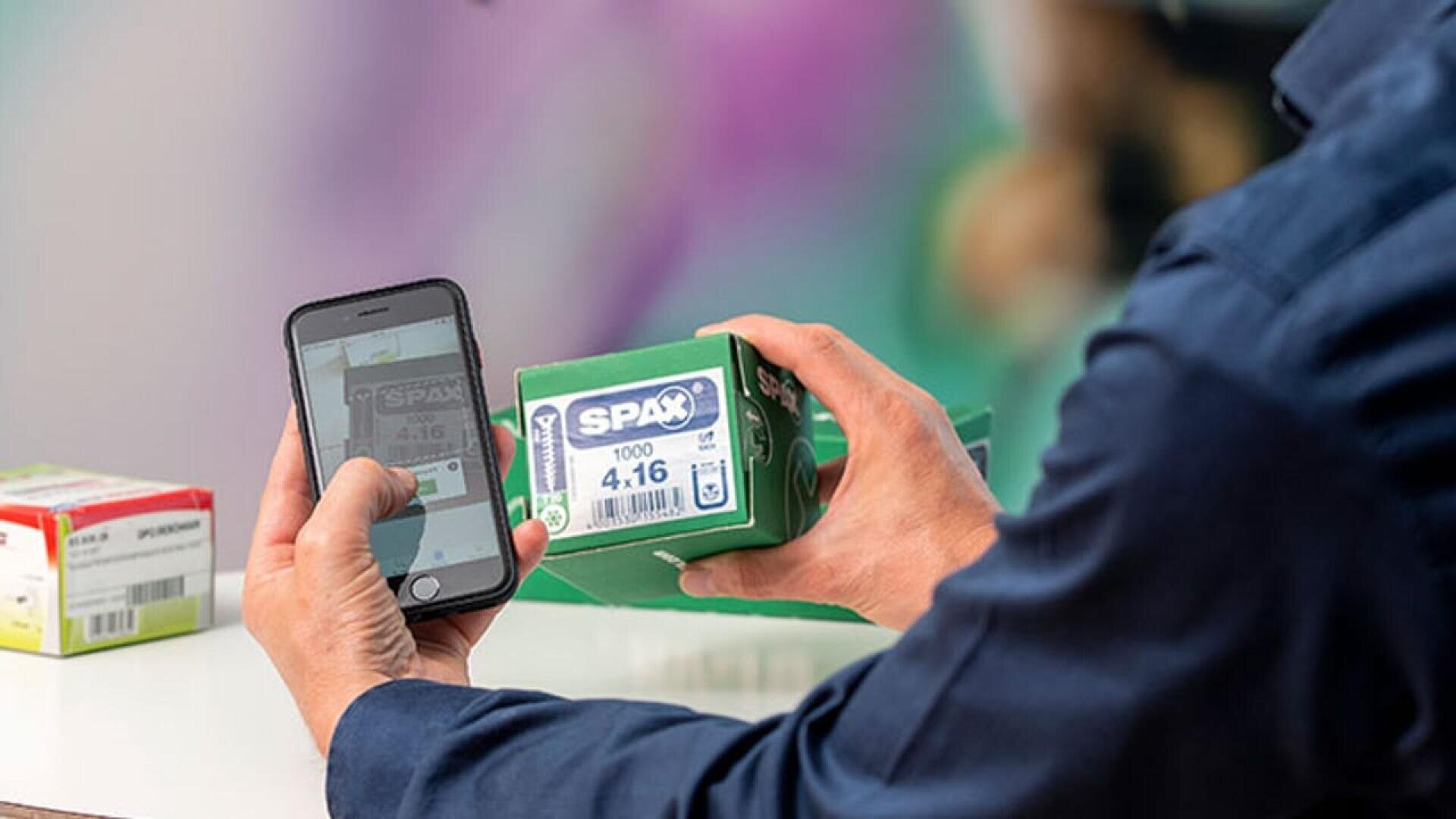 Une personne tient une boîte verte de SPAX et scanne son code-barres ou son étiquette à l'aide d'un smartphone. Une autre boîte est visible sur la table. L'arrière-plan est flou.
