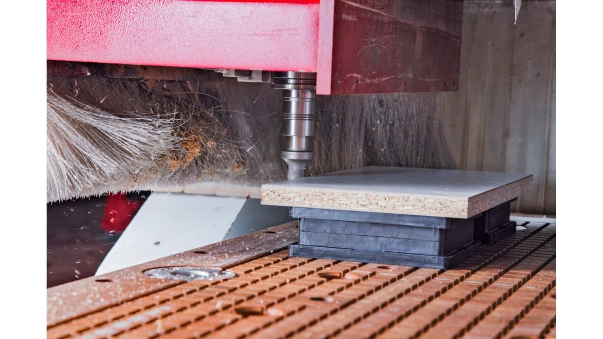 La broche d'une machine CNC découpe ou fraise une feuille de panneau de particules reposant sur des supports. Un capot de machine rouge et une brosse à poussière sont visibles à l'arrière-plan.