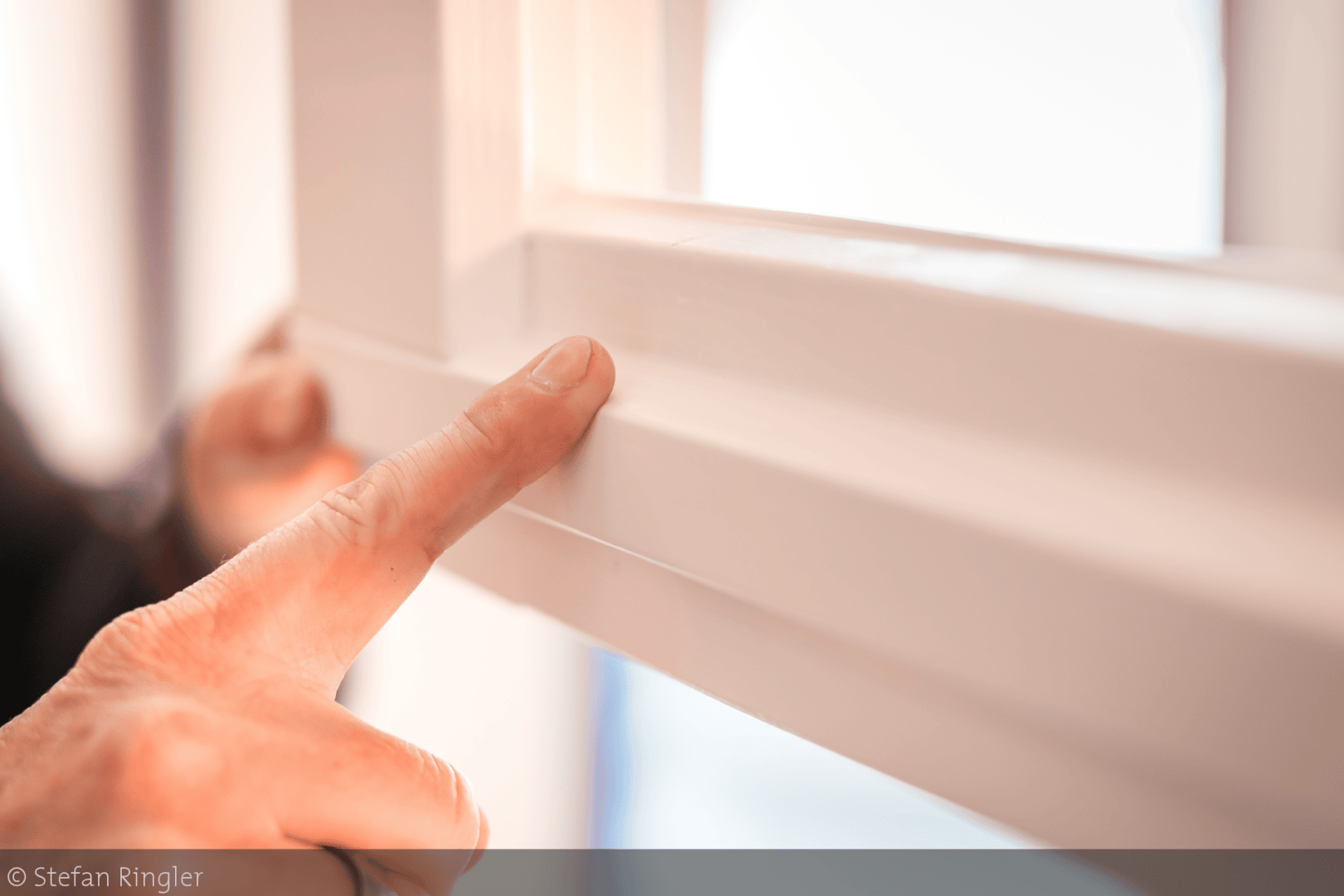 Der Finger einer Person zeigt genau auf den weißen, glatten Rahmen eines Fensters und hebt dessen Oberfläche und Verarbeitung hervor. Das Bild ist auf die Fingerspitze und den Fensterrahmen fokussiert.