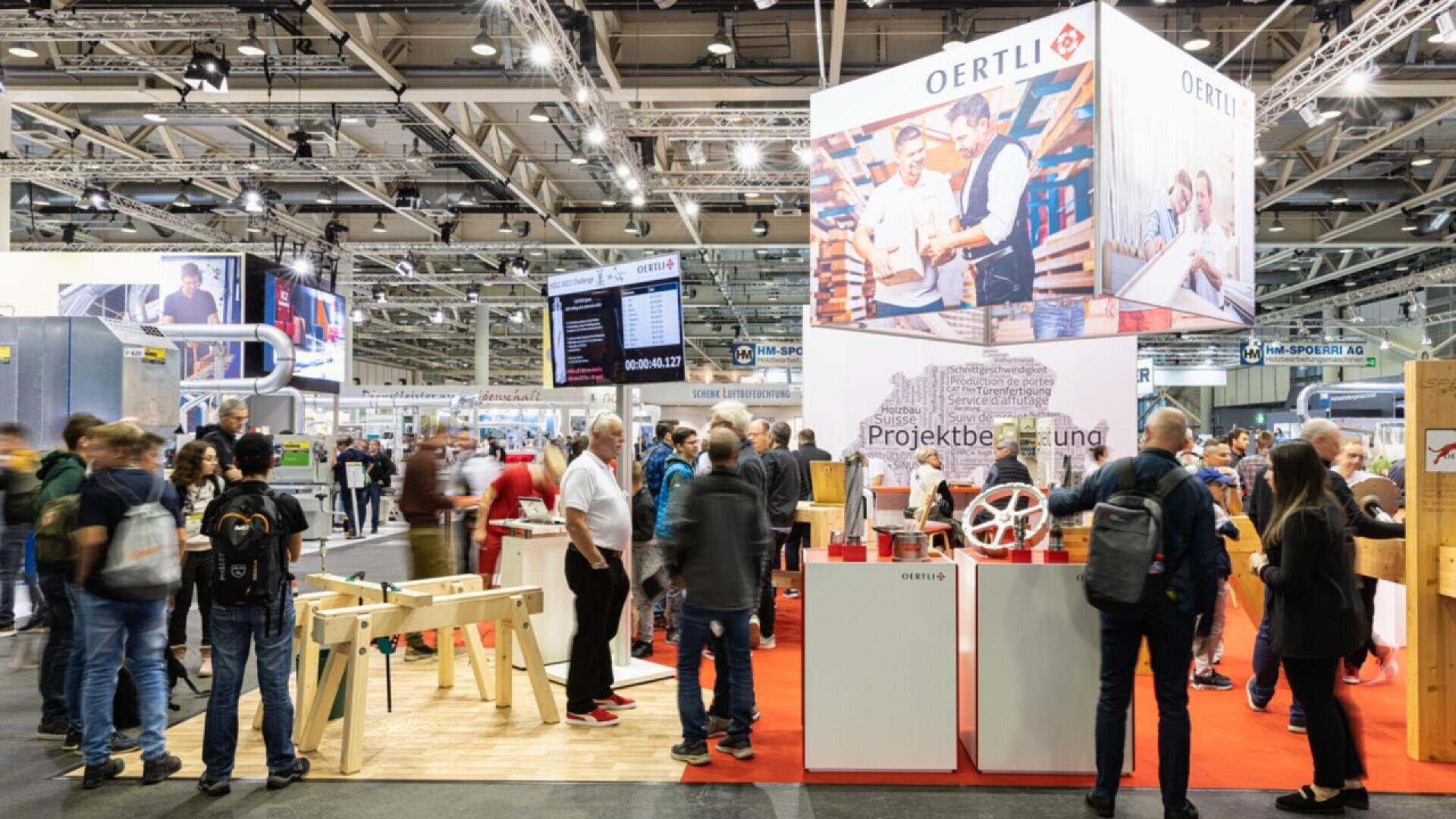 Un stand d'exposition très fréquenté avec la marque OERTLI, des gens qui interagissent et des outils de travail du bois. Le stand comporte des panneaux, des photos et un tapis rouge, dans un grand hall de convention bien éclairé.