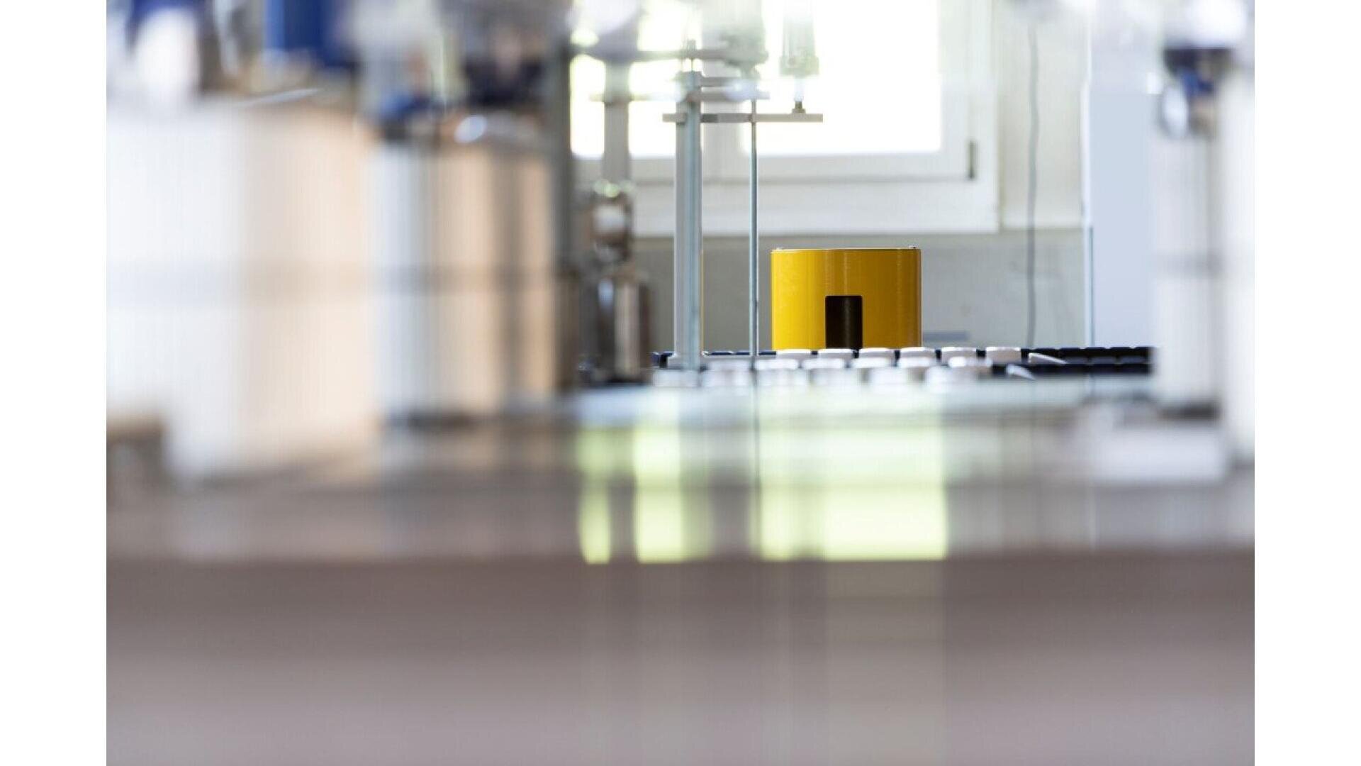 Un objet cylindrique jaune est posé sur une surface réfléchissante dans un laboratoire moderne et lumineux, entouré d'équipements scientifiques flous au premier et à l'arrière-plan.