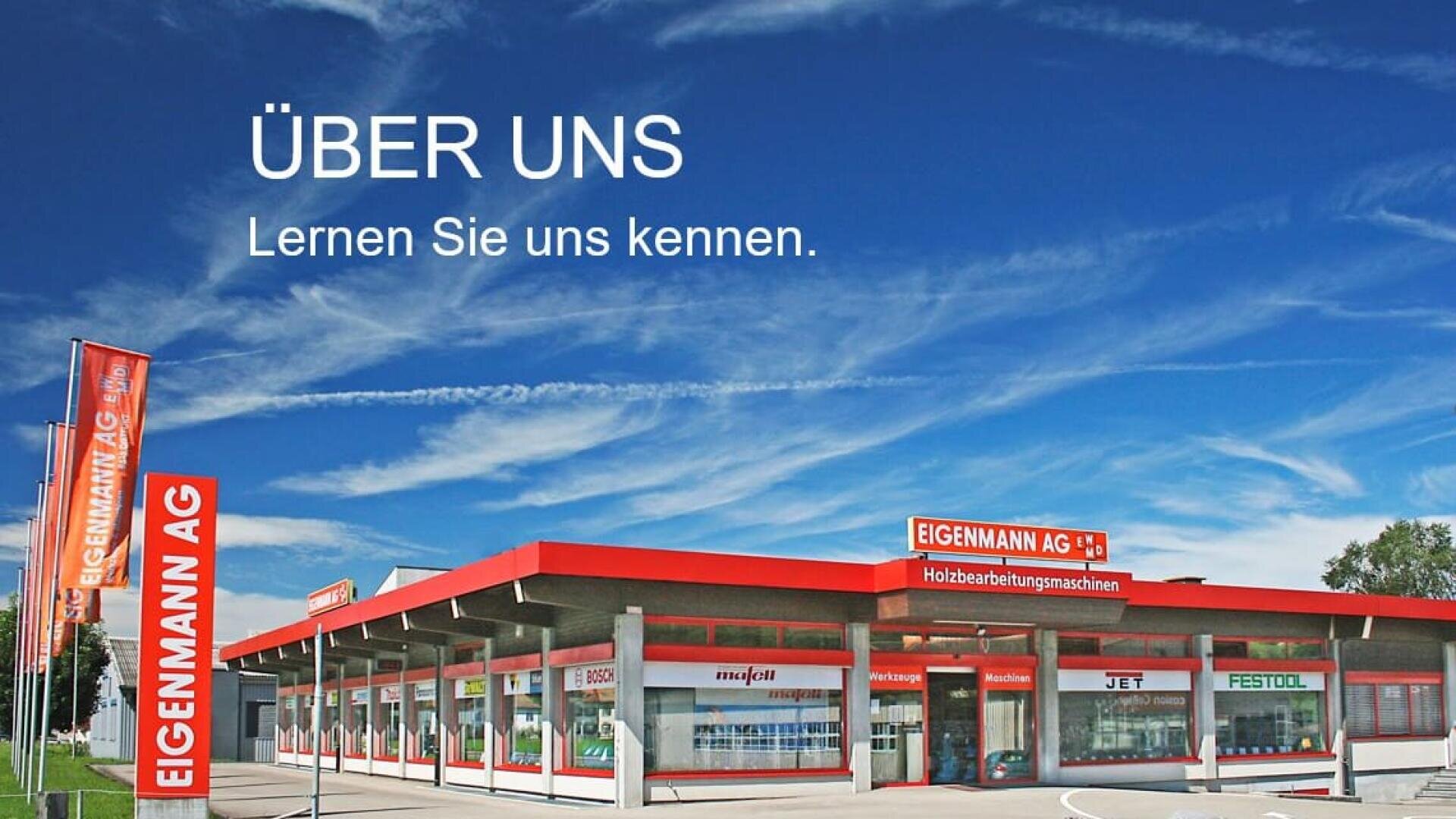Un bâtiment moderne de la société Eigenmann AG avec des accents rouges et des enseignes sous un ciel bleu, avec le texte allemand Über uns. Lernen Sie uns kennen. Les drapeaux indiquent Eigenmann AG et le bâtiment fait de la publicité pour des marques de machines à travailler le bois.