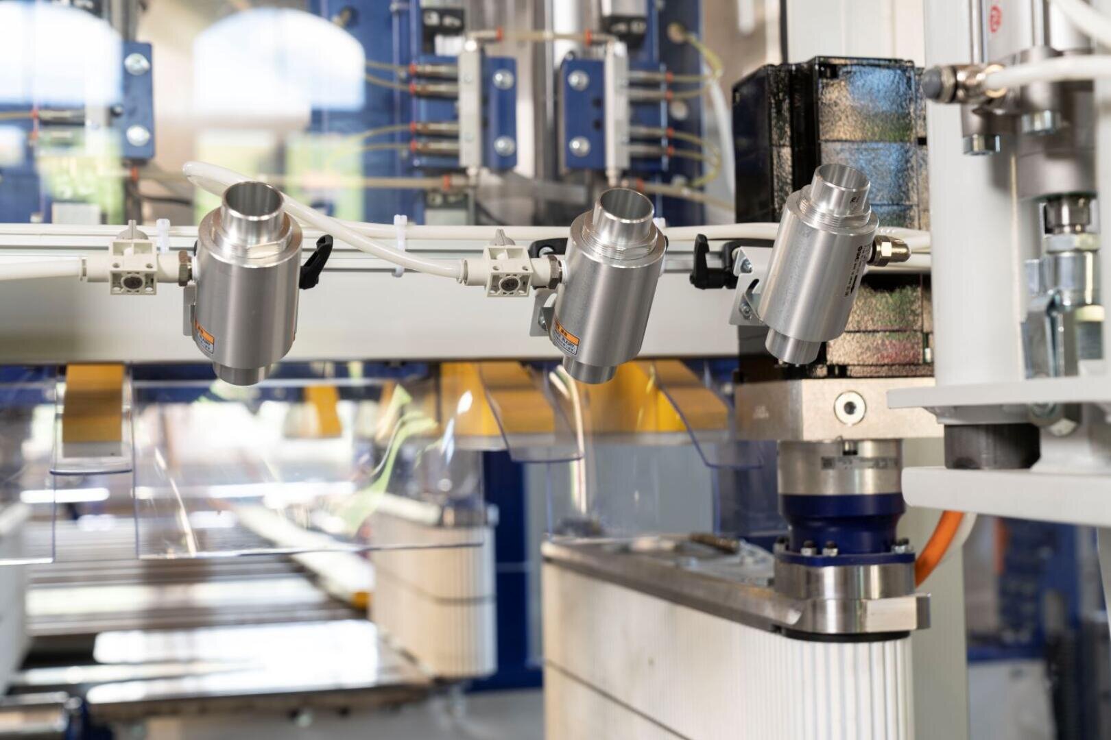 Nahaufnahme einer automatisierten Industriemaschine mit auf einer Schiene montierten Metallsensoren oder Kameras, Verkabelung und durchsichtigen Kunststofffolien, die in einer Fabrik oder Fertigungsumgebung eingesetzt wird.