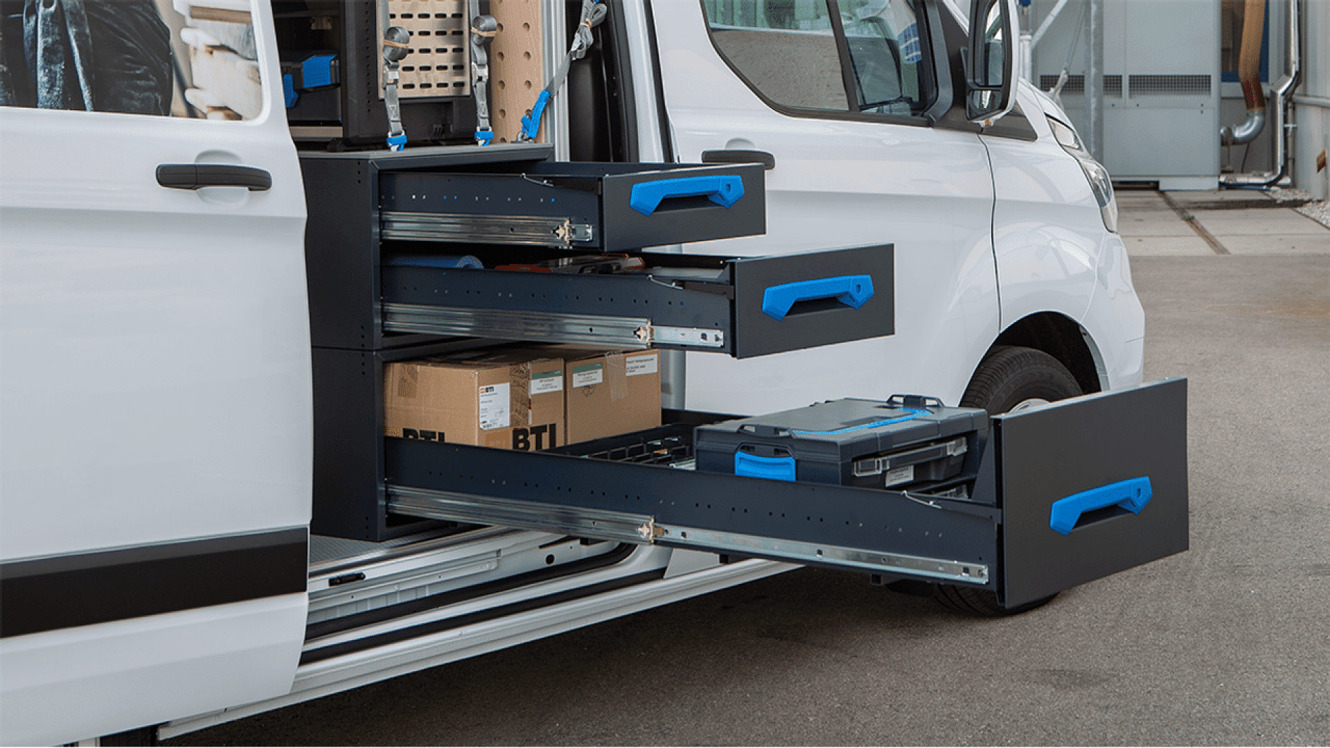 Une camionnette blanche dont la porte latérale est ouverte, montrant plusieurs tiroirs de rangement noirs avec des poignées bleues tirées, contenant des boîtes et des outils organisés pour un accès facile.