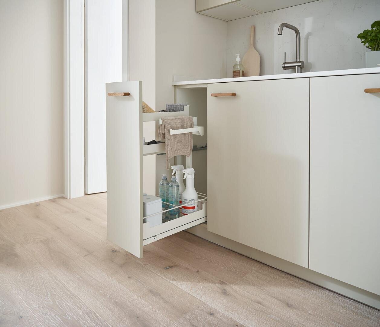 Eine moderne Küche mit hellem Holzboden verfügt über einen ausziehbaren Schrank unter der Spüle, in dem Reinigungsmittel, Sprühflaschen, Schwämme und Handtücher untergebracht sind. Das Design ist minimalistisch und aufgeräumt.