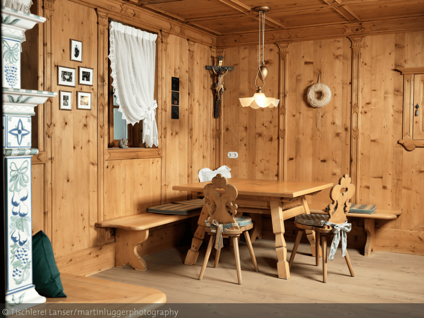 Eine gemütliche Essecke aus Holz mit geschnitzten Stühlen, einer Bank und einem quadratischen Tisch. Der Raum hat holzgetäfelte Wände, ein Fenster mit einem Spitzenvorhang, gerahmte Bilder, ein Kruzifix und eine Hängelampe.