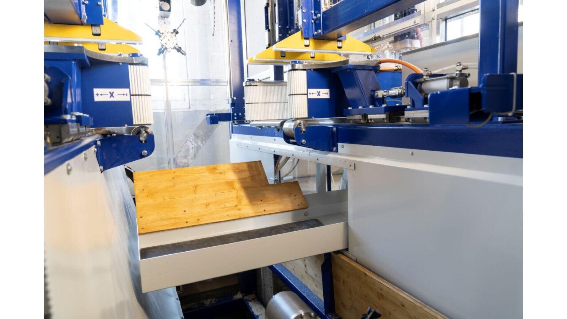 Vue rapprochée d'une machine industrielle dans une usine, avec des pièces métalliques bleues et jaunes, des panneaux de commande et un tapis roulant sur lequel est posée une planche de bois inclinée.