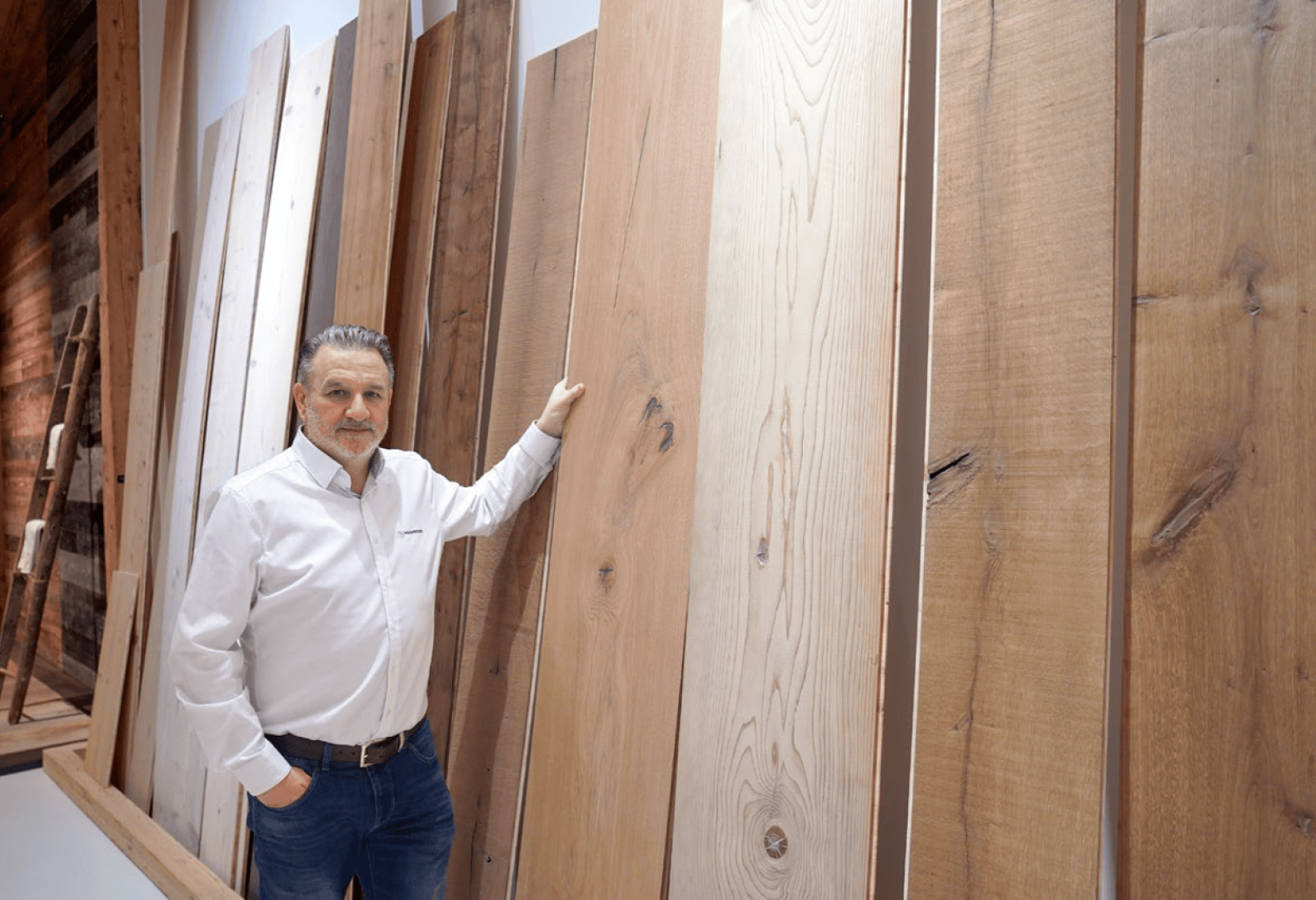 Ein Mann in einem weißen Hemd steht in einem Innenraum und berührt ein großes Holzbrett inmitten mehrerer aufrecht stehender Holzplatten, die an einer Wand angebracht sind.