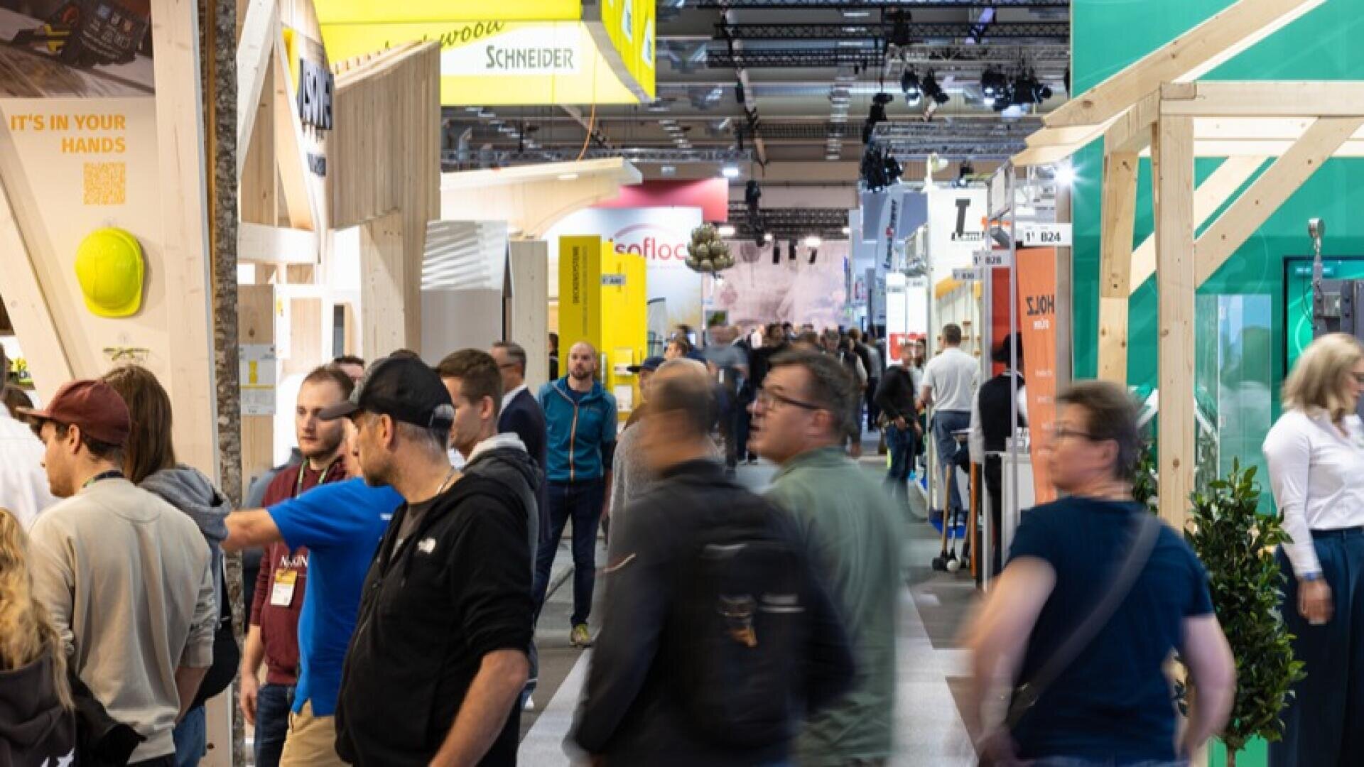 Un salon professionnel ou une exposition en intérieur très fréquenté, avec des gens qui se promènent et parcourent divers stands et présentoirs colorés. Certains visiteurs discutent, tandis que d'autres regardent les produits ou les enseignes. L'atmosphère est animée et bondée.
