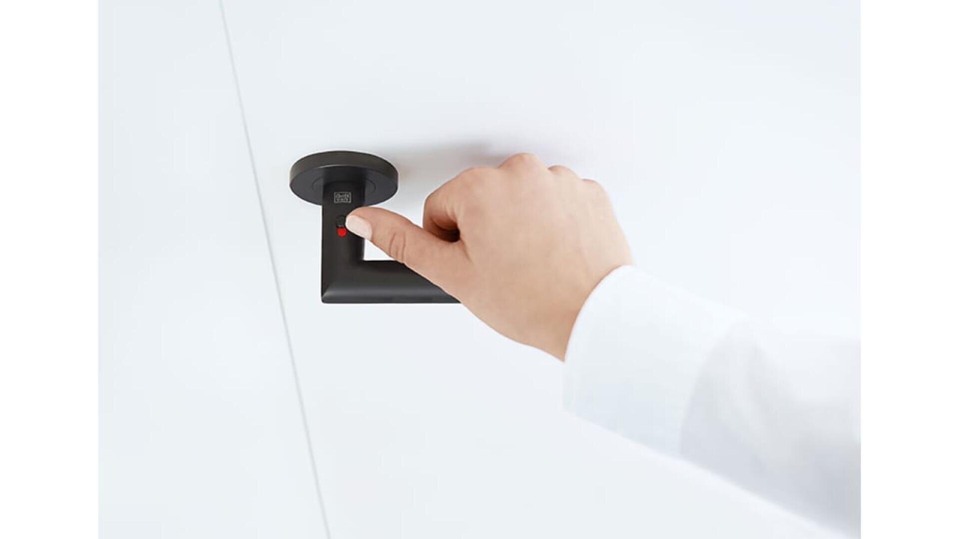 Une personne vêtue d'une manche blanche appuie son pouce sur un lecteur d'empreintes digitales situé sur une poignée de porte moderne noire sur fond blanc.