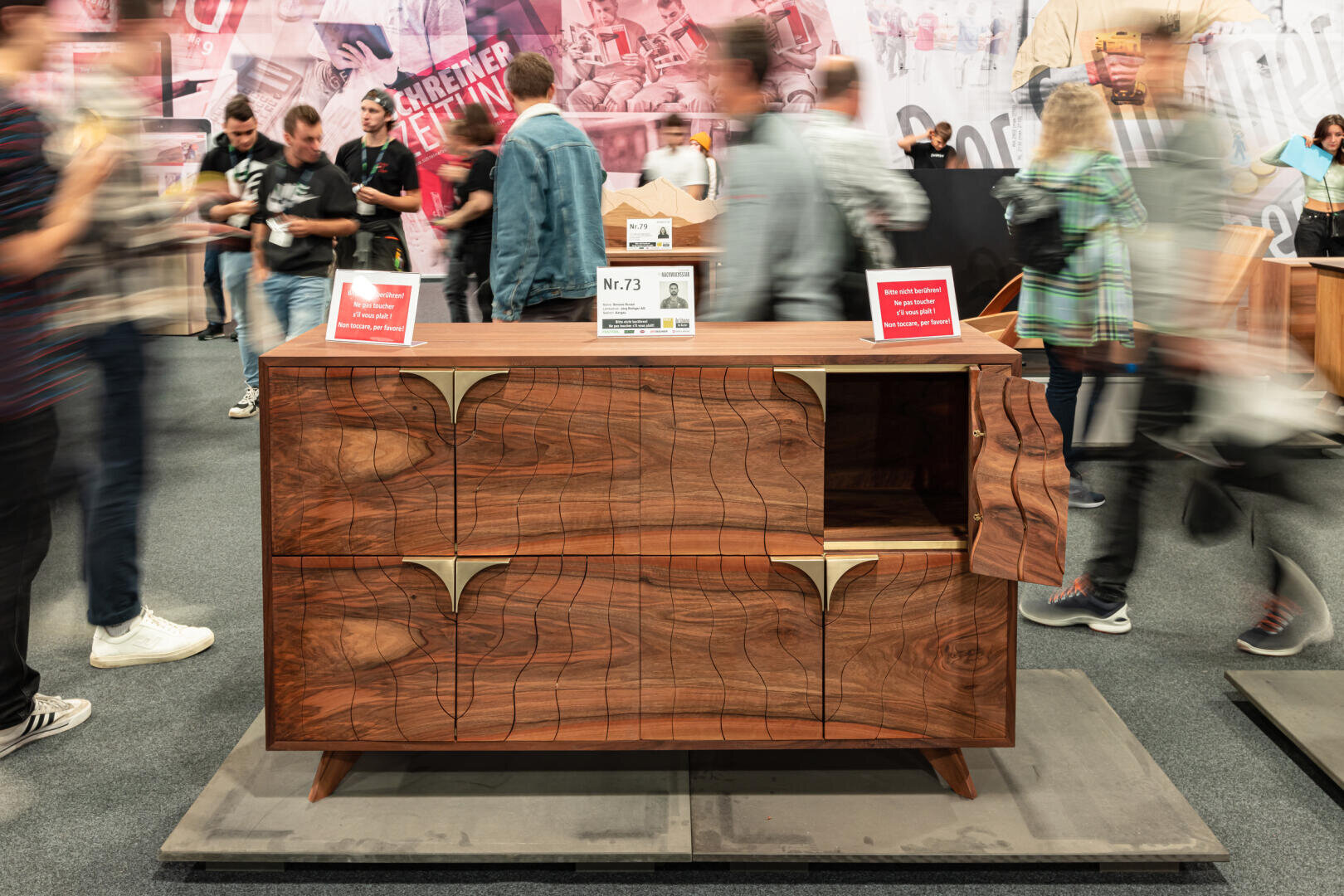 Une armoire moderne en bois avec des motifs de grain complexes et des accents en laiton est présentée lors d'une exposition, entourée d'un mouvement flou de personnes passant devant. Des panneaux et des cartes d'information sont placés au-dessus de l'armoire.