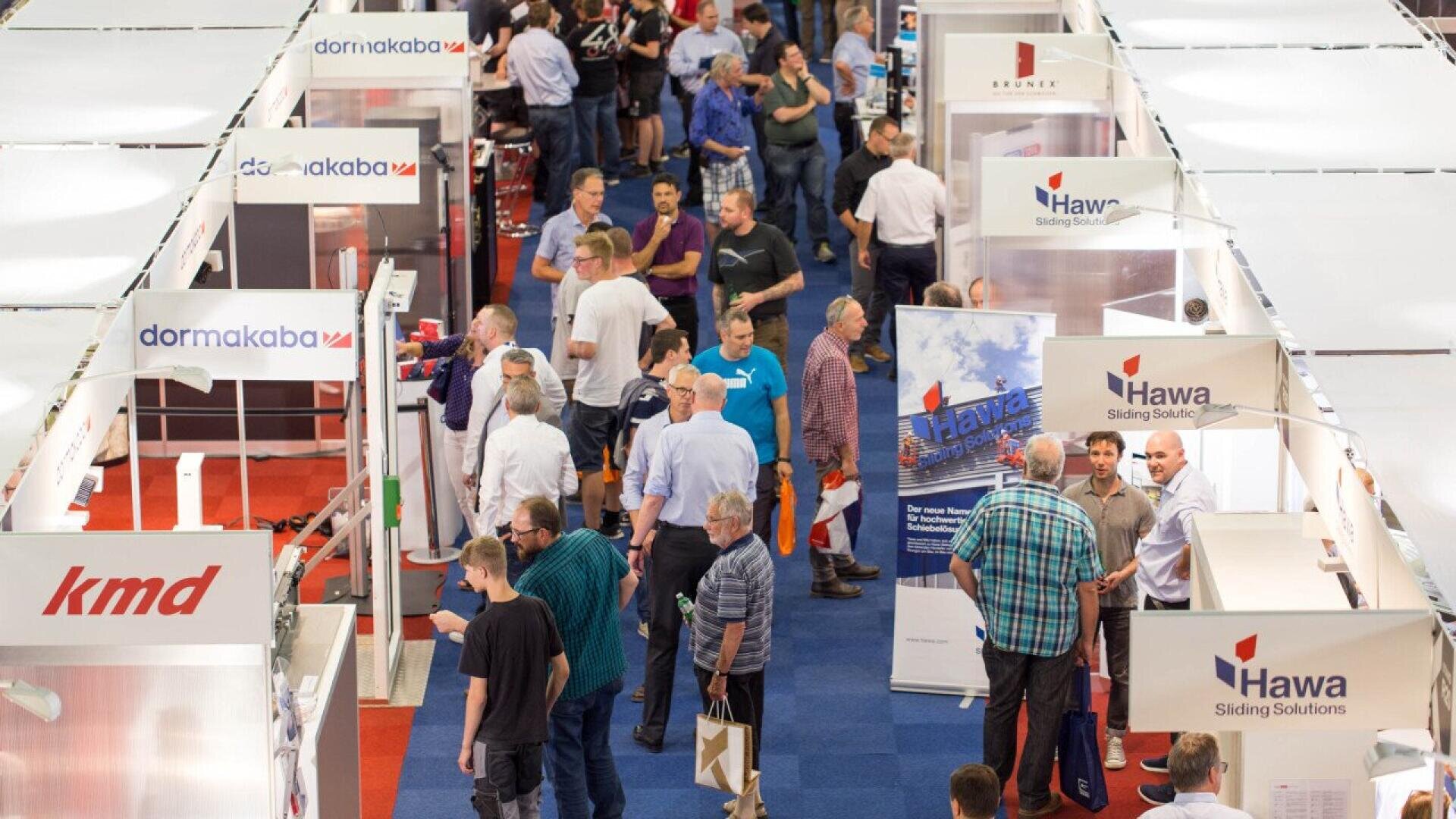 Un salon professionnel intérieur très fréquenté, avec de nombreuses personnes circulant entre les stands présentant des enseignes, des bannières et des produits de l'entreprise sous des éclairages suspendus blancs. Le sol est recouvert de dalles de moquette bleues et rouges.