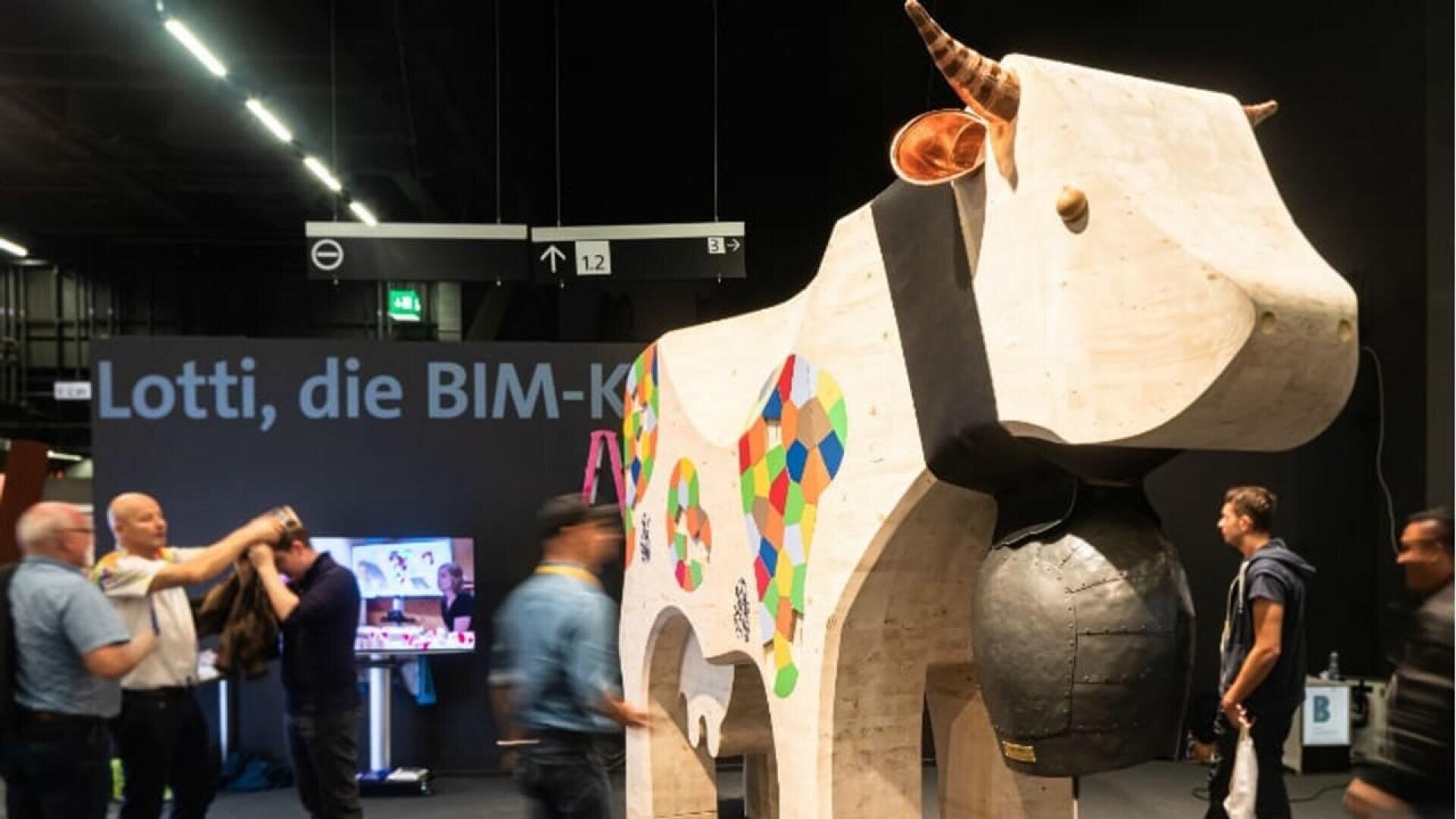 Une grande sculpture de vache colorée avec des cornes et un pis noir est exposée à l'intérieur. Plusieurs personnes sont rassemblées autour d'elle, certaines prenant des photos. Un panneau sur le mur indique Lotti, die BIM-K.