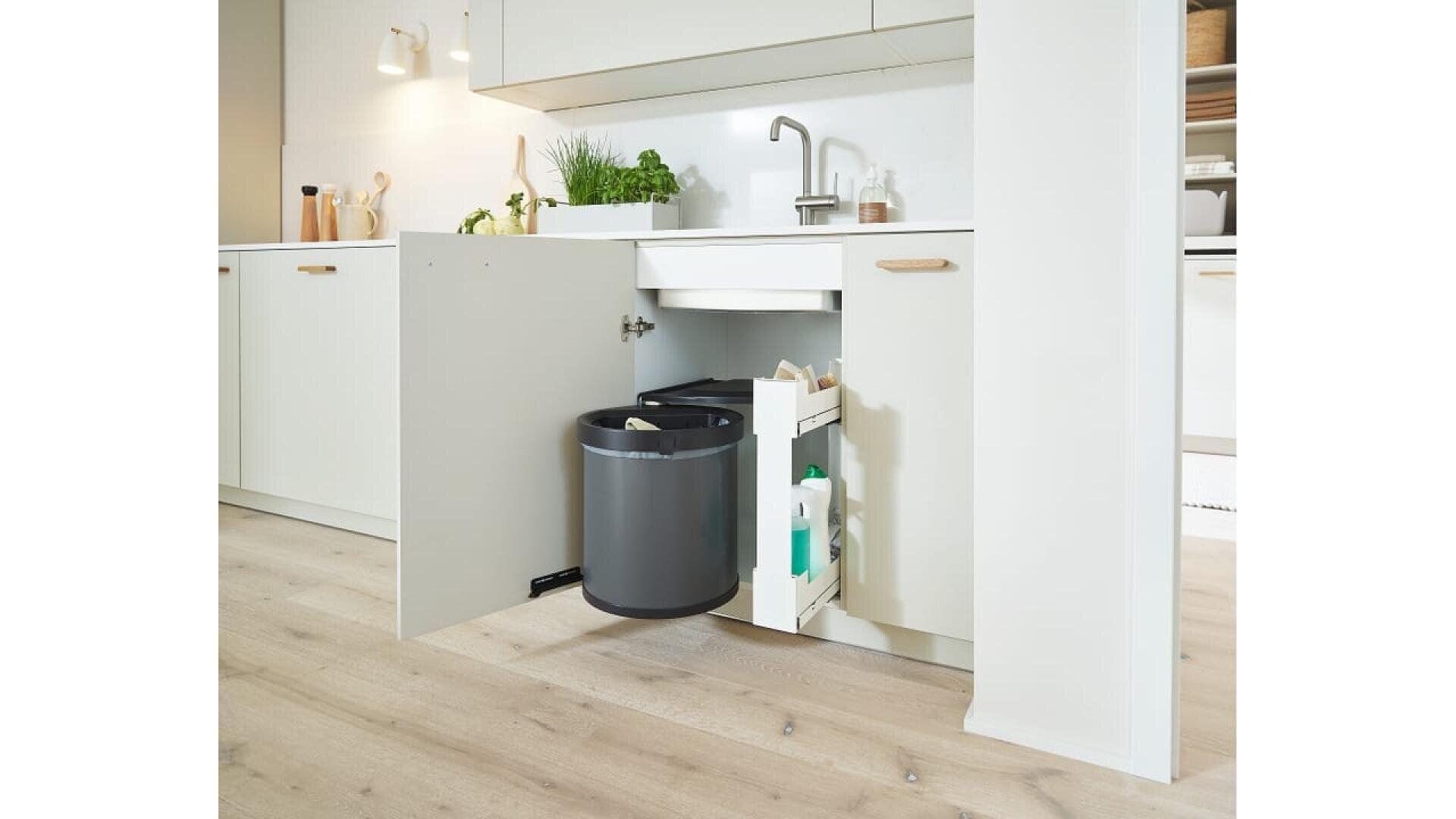 Un meuble de cuisine ouvert sous l'évier révèle une poubelle ronde grise et un organisateur extractible contenant des produits de nettoyage. La cuisine a des armoires de couleur claire, un sol en bois et des plantes en pot sur le comptoir.