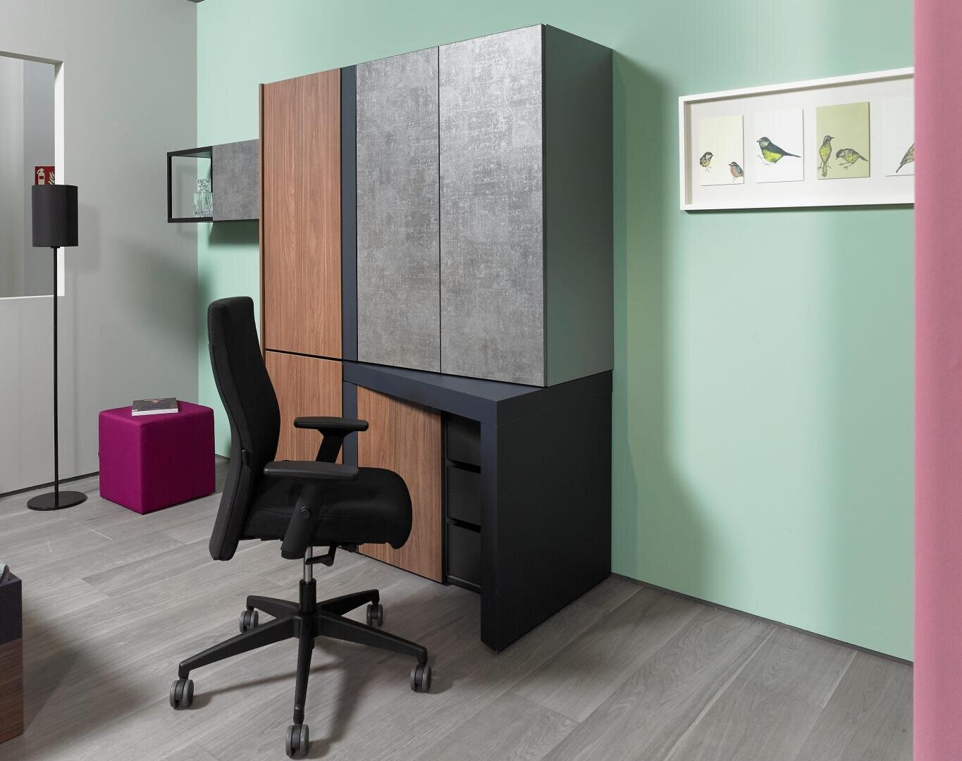 Un bureau moderne avec une chaise ergonomique noire, des armoires en bois et grises, des murs vert clair, un sol gris, un ottoman magenta, un lampadaire et des illustrations d'oiseaux encadrées au mur.