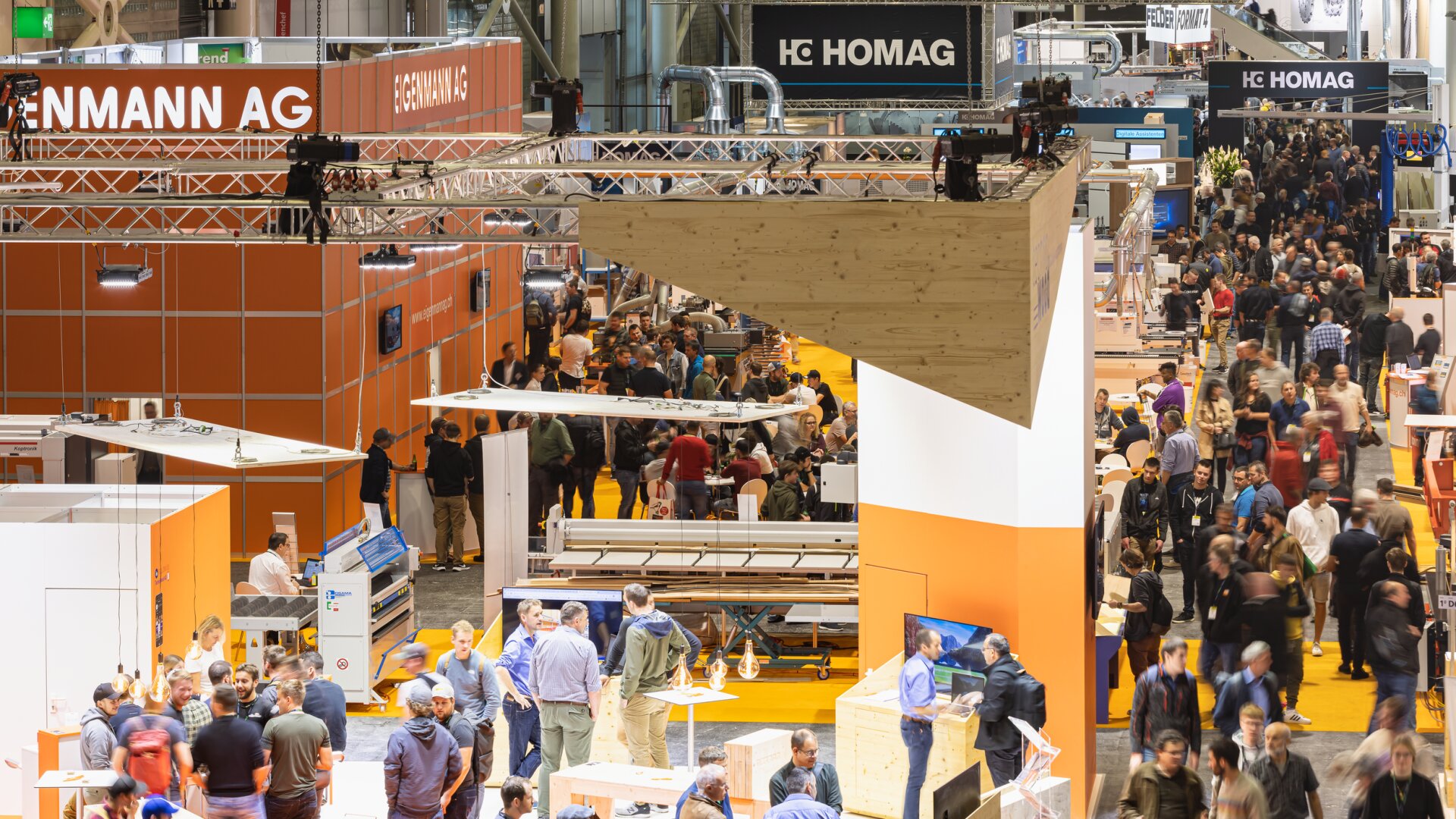 Le sol d'un salon professionnel est encombré de stands orange et blancs, de divers équipements et de groupes de personnes qui marchent et discutent. Des enseignes de marques telles que Homag et Henmann AG sont visibles au-dessus de cette scène animée.