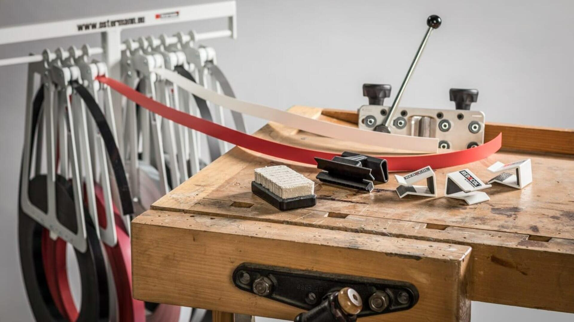 Un établi en bois avec des outils d'affûtage, des pinces, des bandes abrasives de différentes couleurs suspendues à un support, et un gabarit d'affûtage placé sur la table. L'ensemble suggère un espace de travail pour l'affûtage des lames ou le travail du bois.